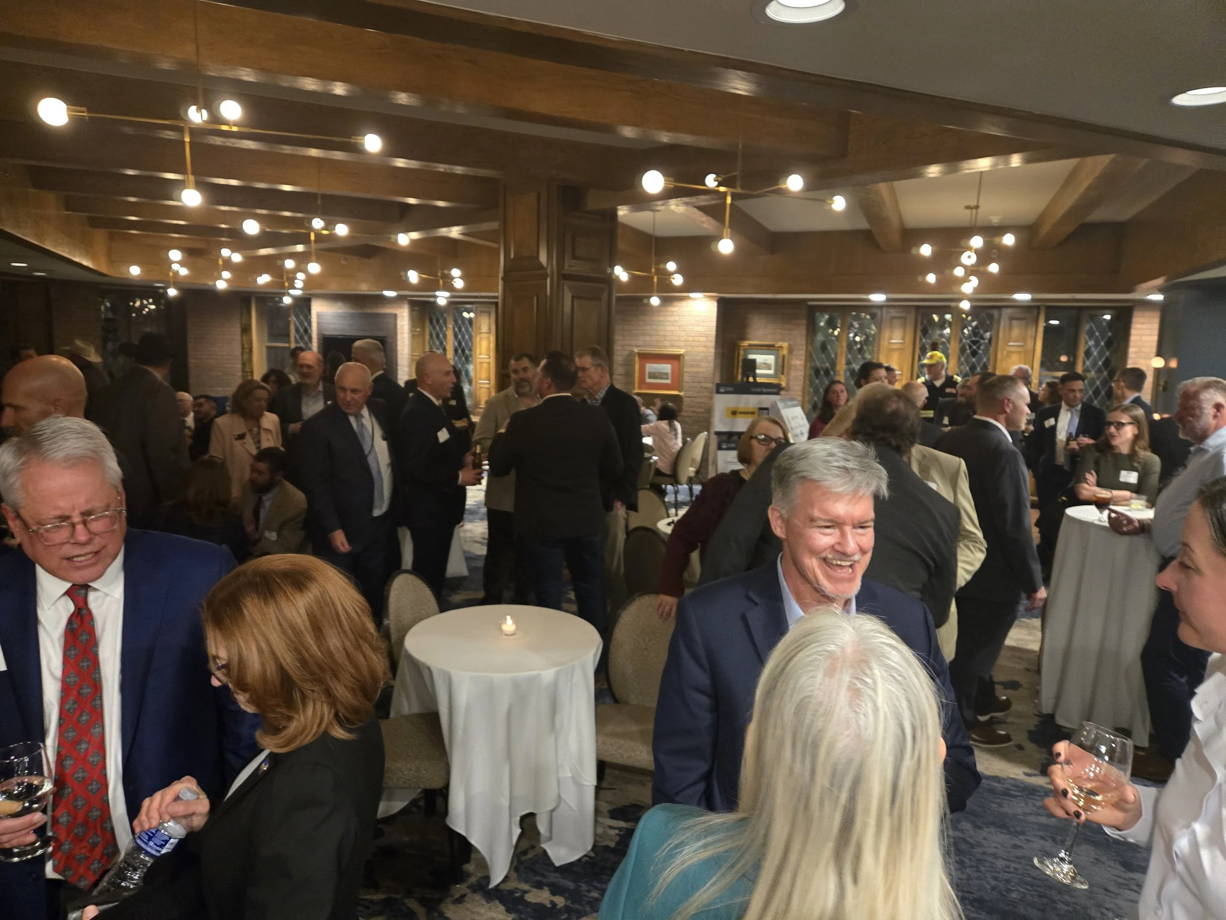 People socializing at a formal event in a decorated dining room or banquet hall, with men and women in business attire, some holding drinks, and a fireplace in the background.