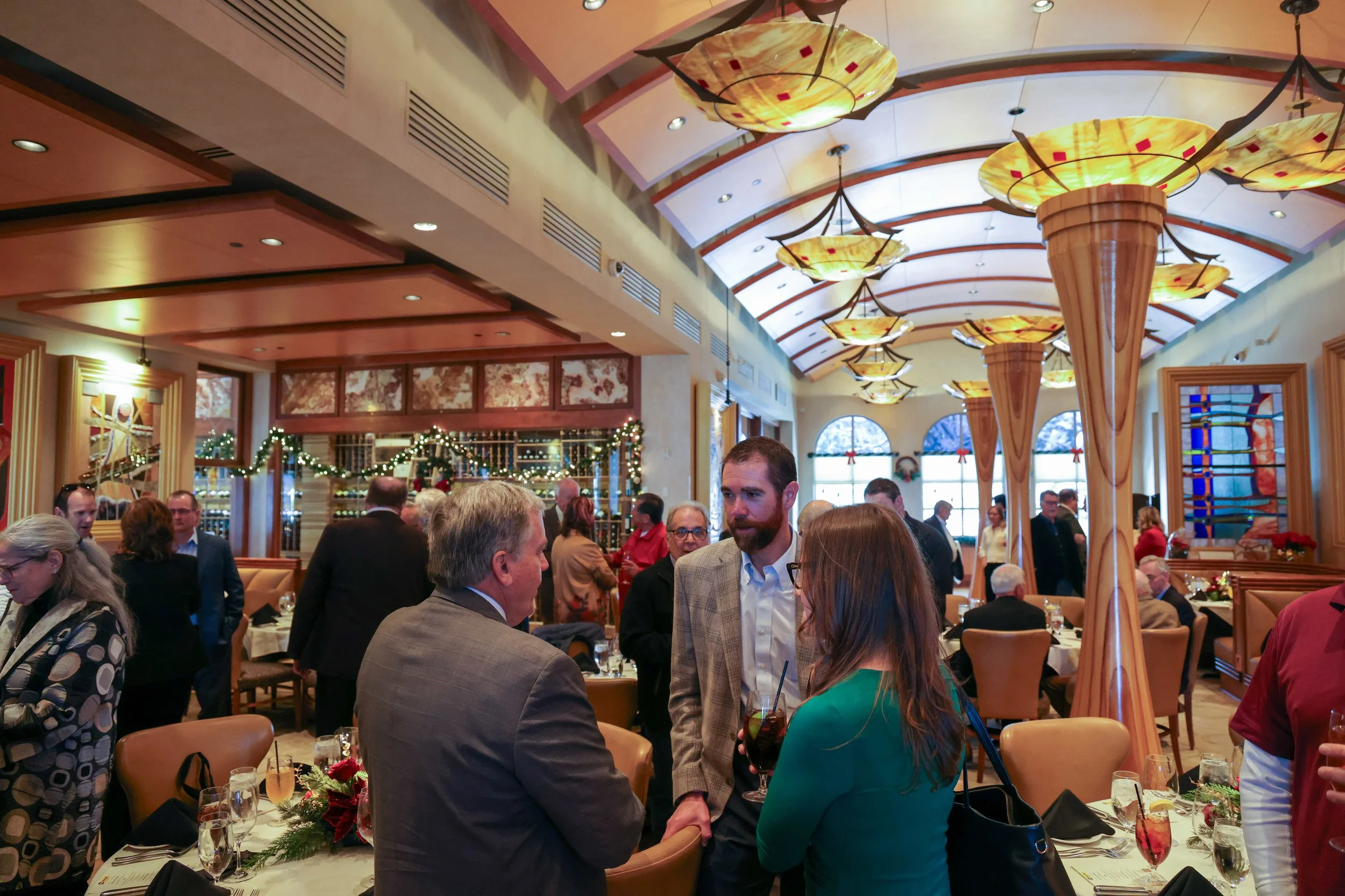 People attending a celebration or holiday event in a decorated restaurant or banquet hall, with festive lights and decorations, and many individuals engaged in conversation.