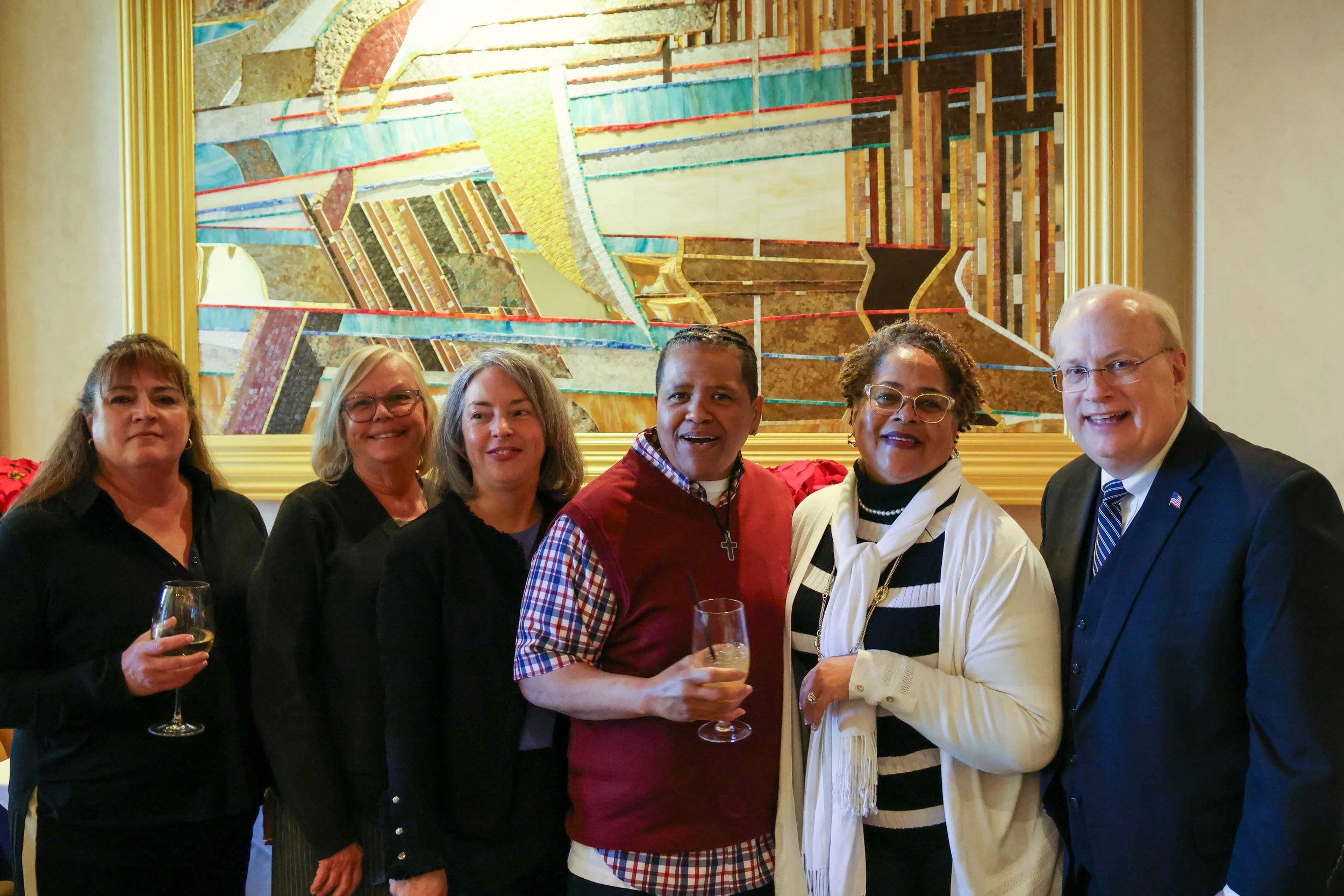 Six people standing together in front of a large, colorful abstract artwork. They are smiling and holding glasses of wine or champagne at a social gathering.
