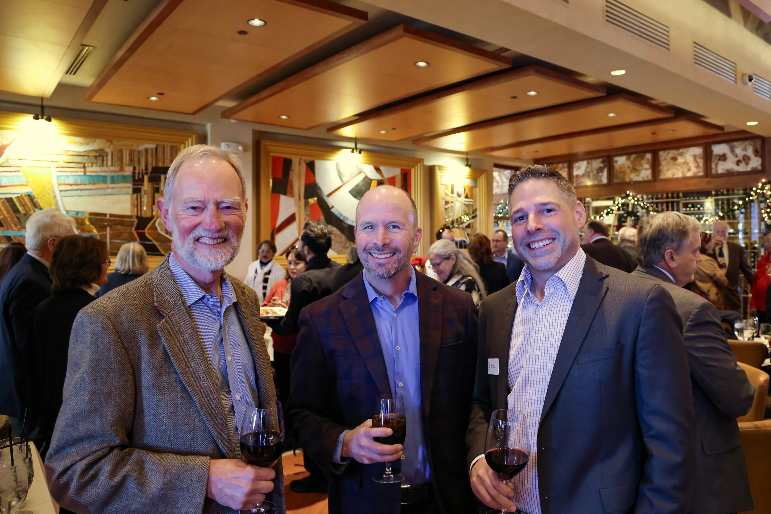 Three men in suits holding glasses of red wine at a crowded indoor event, smiling for the camera