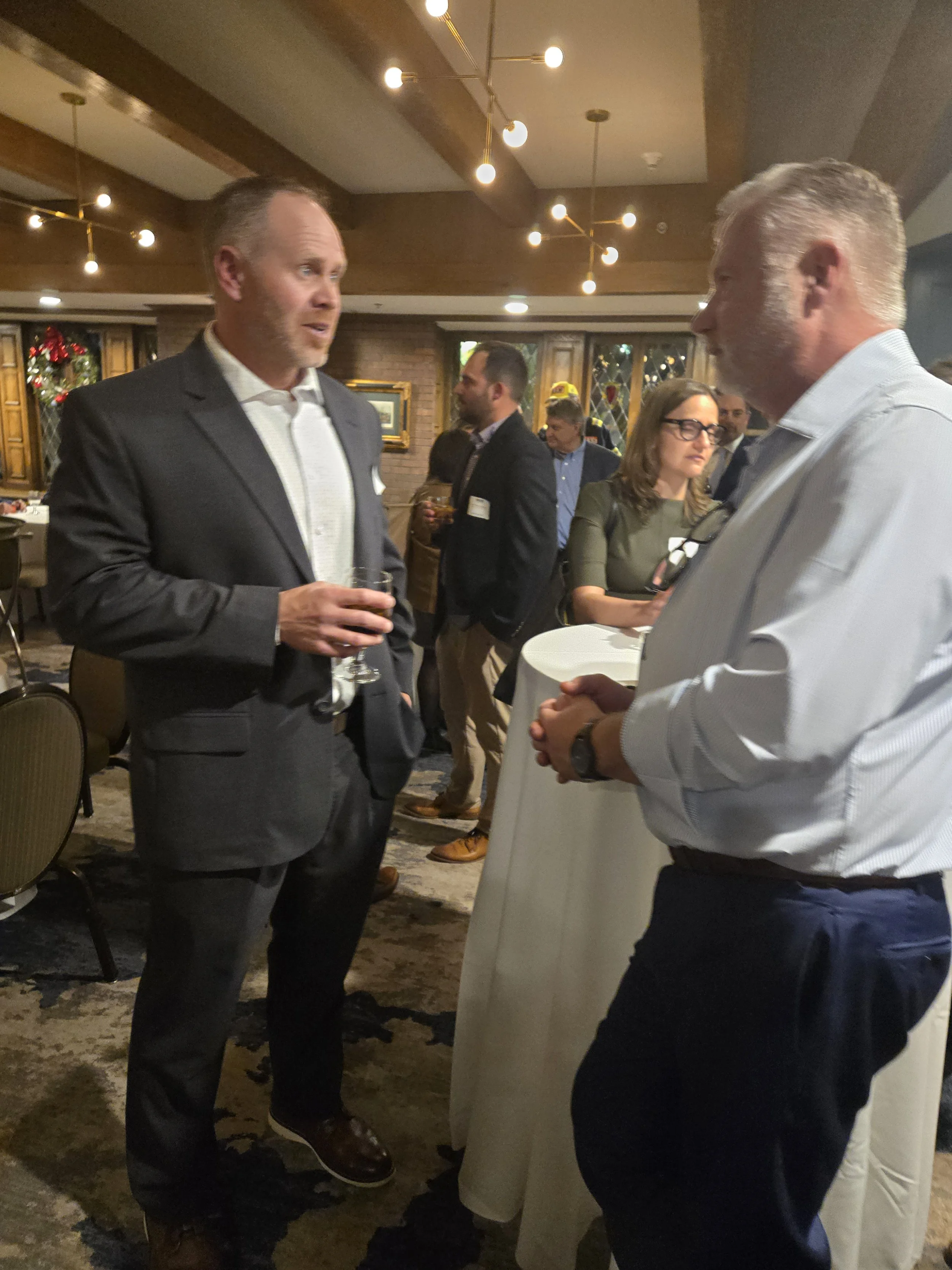 Two men are engaged in conversation at a networking event in a decorated, warmly lit room, with other attendees in the background.