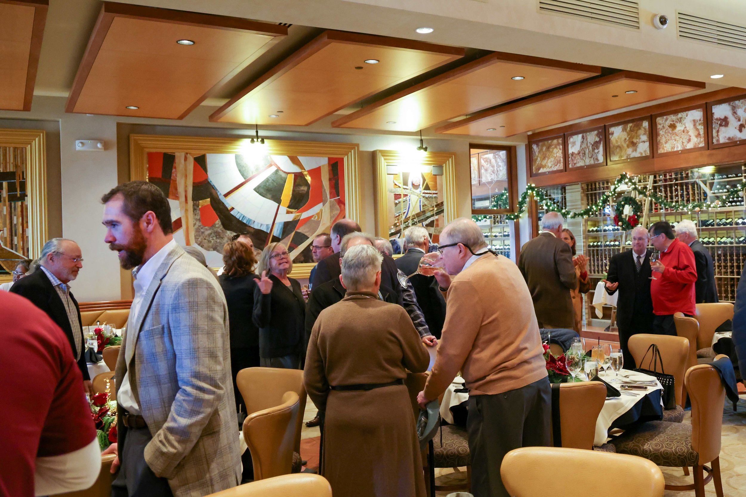 A group of people gathered in a festive restaurant or banquet hall with holiday decorations, including garlands and wreaths, and wine racks in the background. The scene depicts socializing and dining during a holiday celebration.