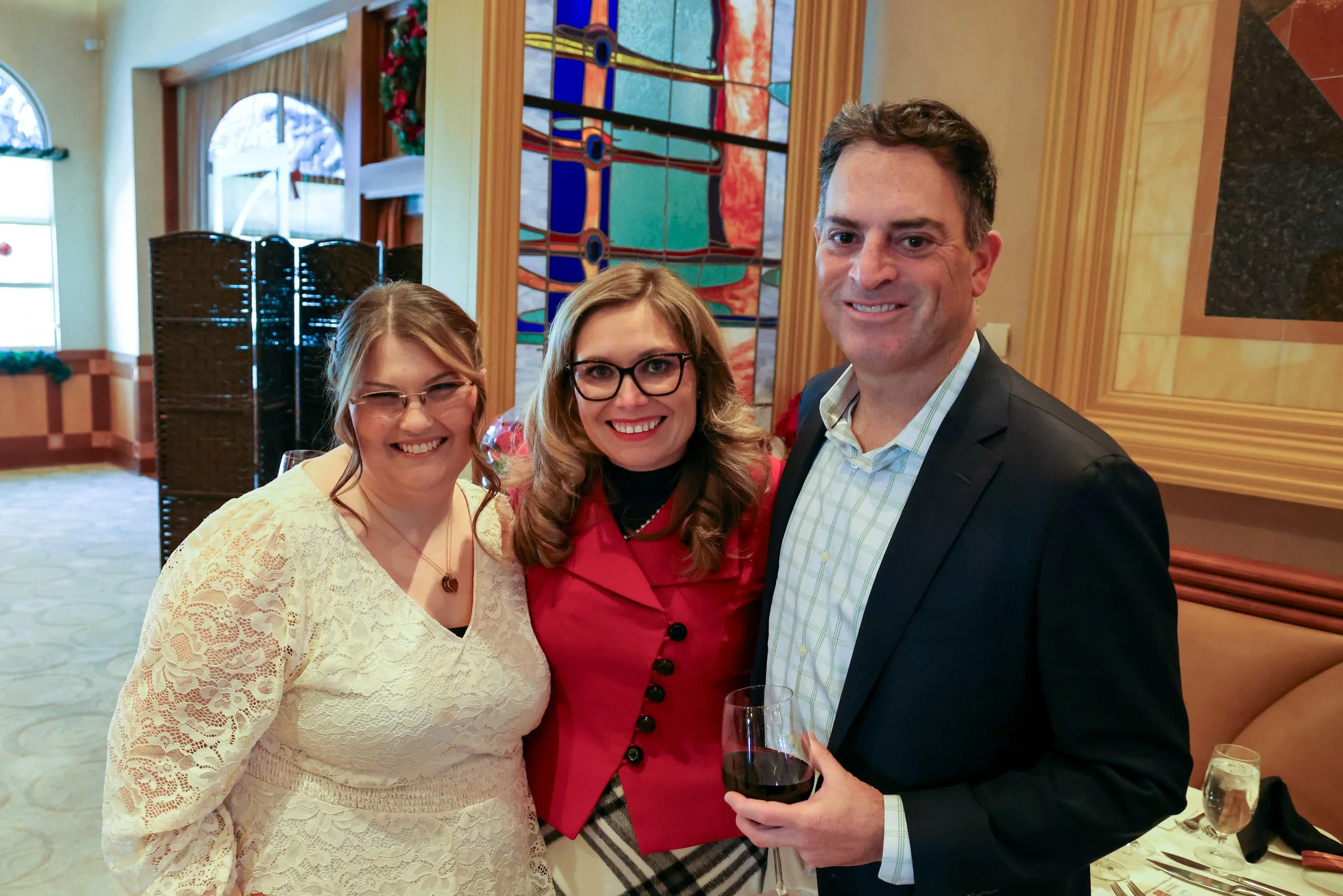 Three people smiling at a social gathering, standing indoors in front of a stained glass window, with one holding a glass of red wine.