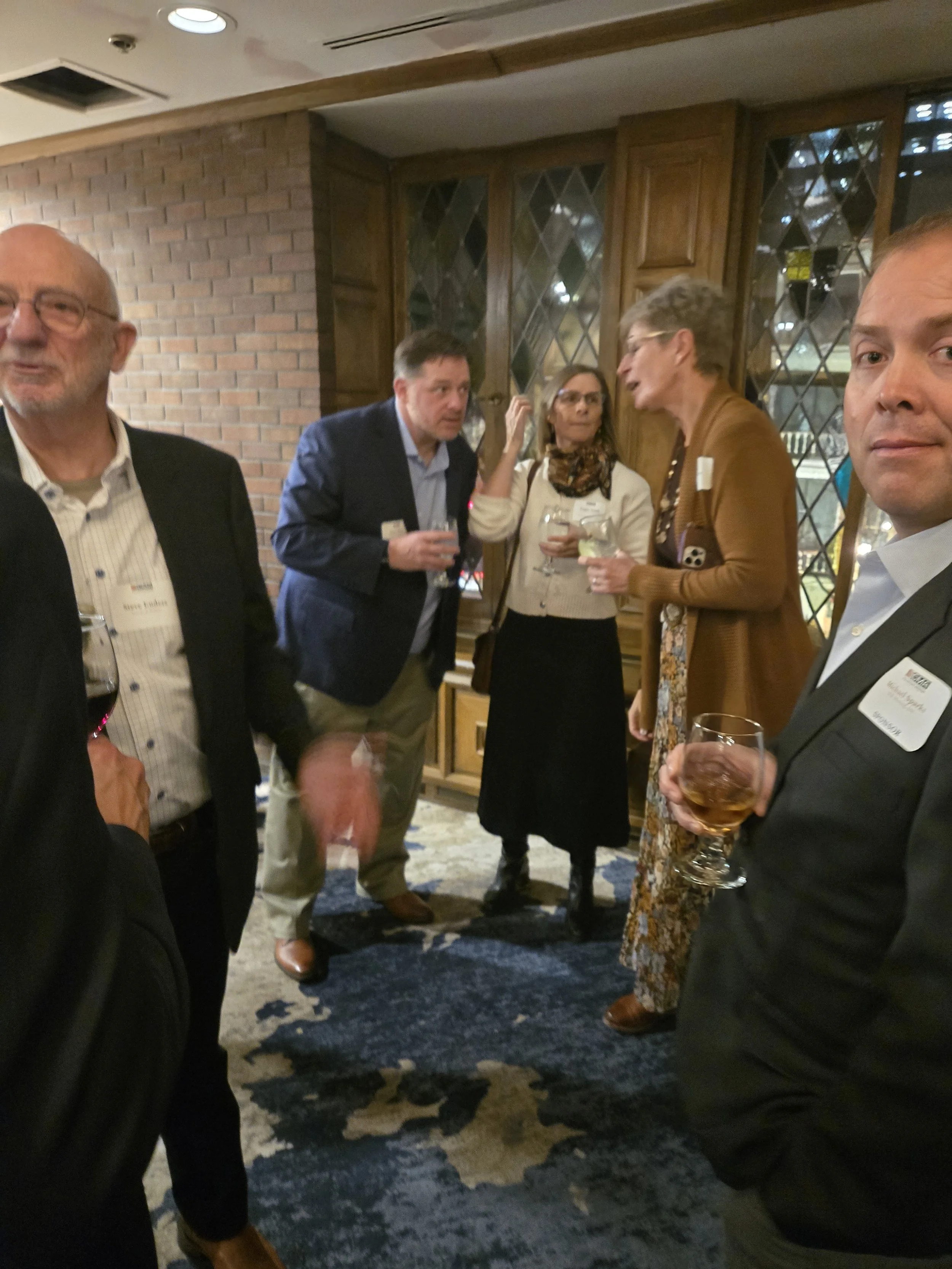 A group of five adults at a social event inside a room with wood-paneled walls and a large window, sipping drinks and engaging in conversation.