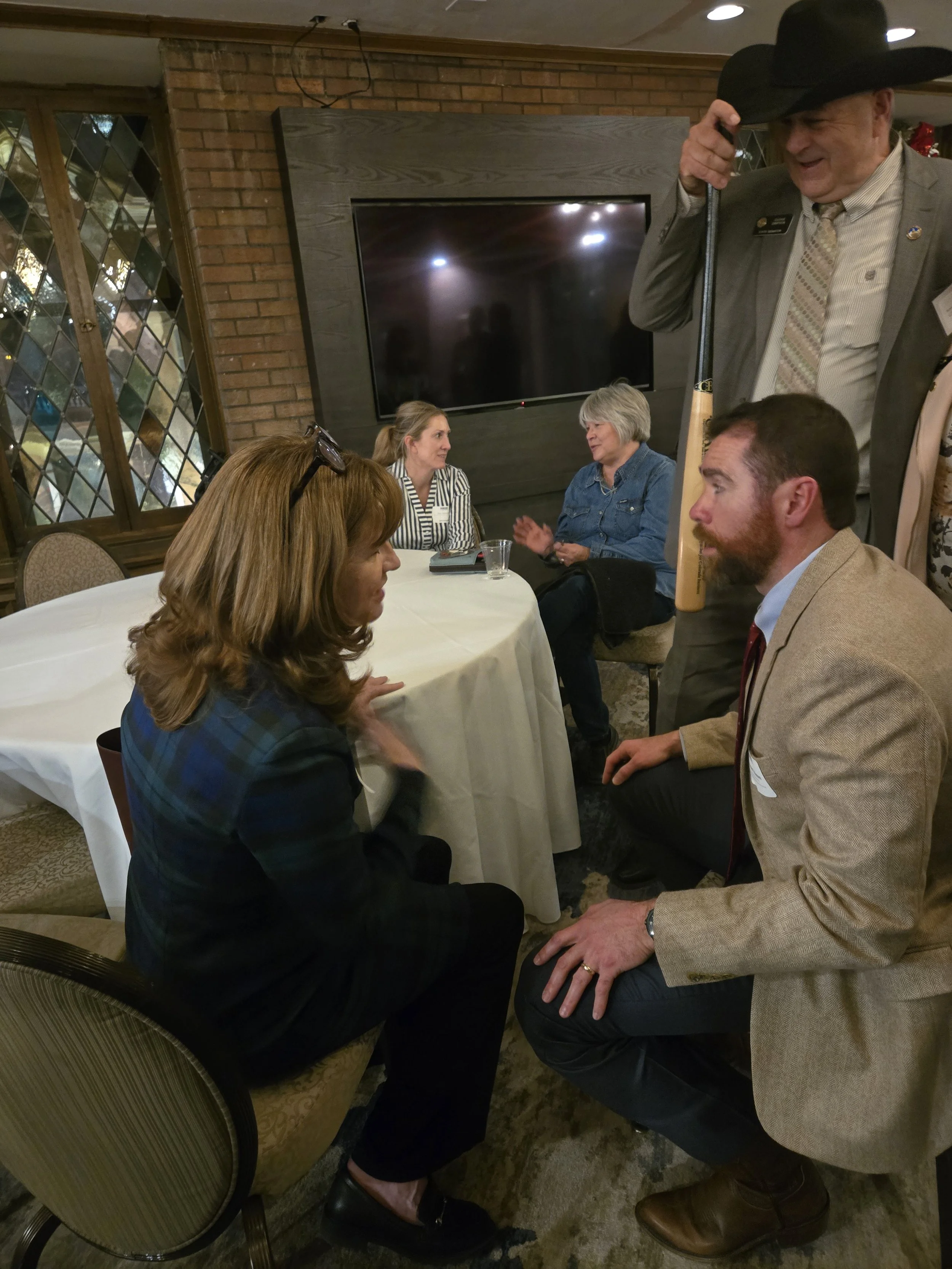 A group of people engaged in conversation at a social gathering in a cozy room. One man is holding a shotgun over his shoulder, and a woman is sitting on a chair with her hand near her face, talking to a man kneeling in front of her. Other people are