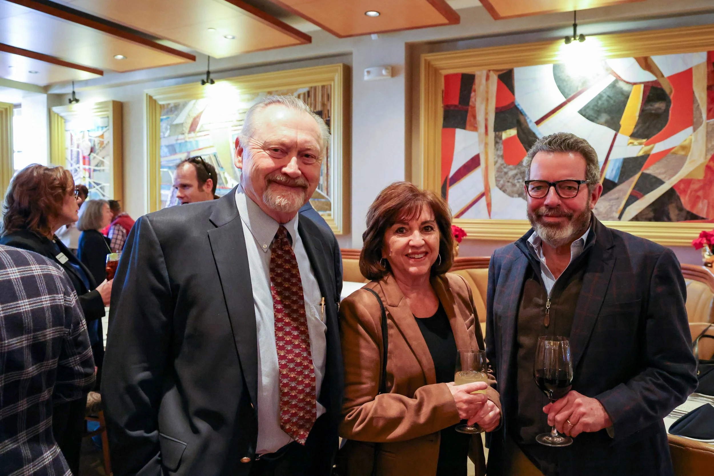 Three adults at a social gathering, smiling and holding drinks, indoors with colorful wall art and other people in the background.