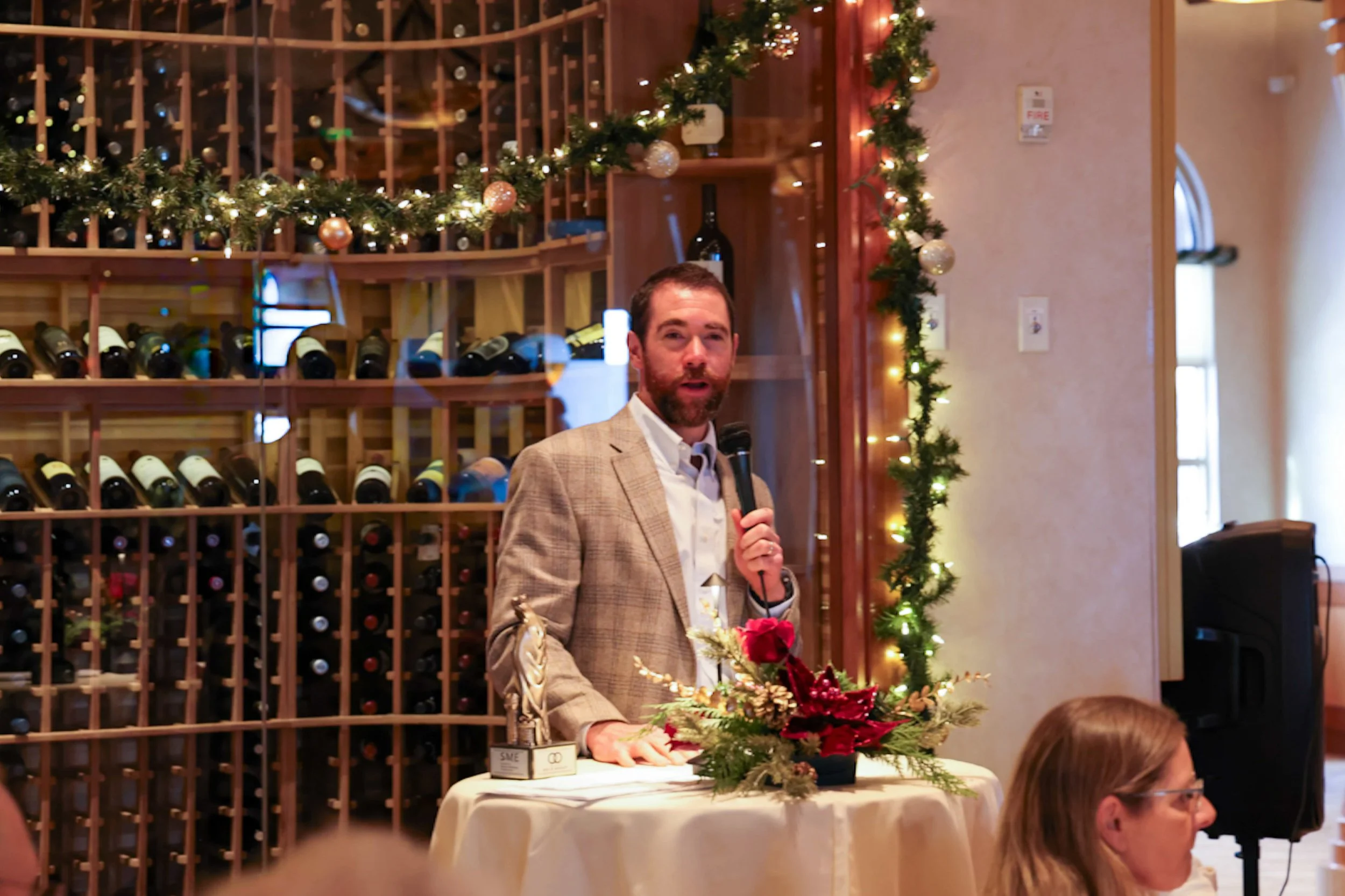 A man in a beige blazer speaking into a microphone at a holiday event, standing behind a table decorated with Christmas flowers, with a wine rack and festive garland in the background.