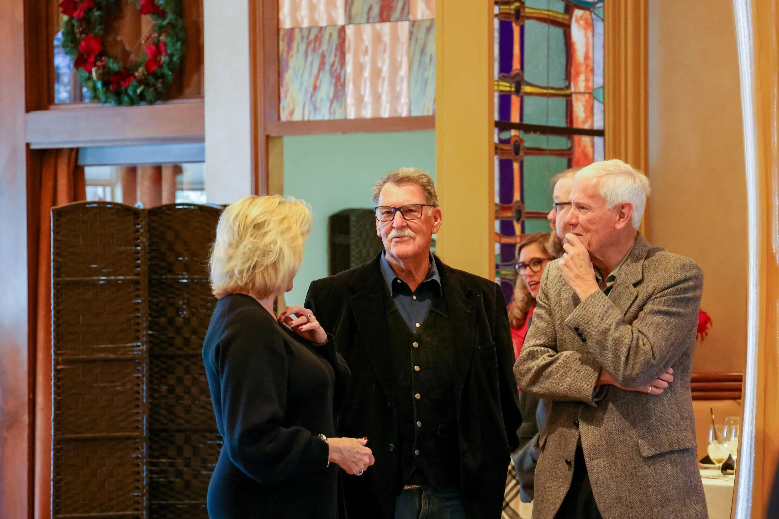 Four older adults in conversation at a gathering, standing in a room with colorful artwork and a wreath on a glass door in the background.
