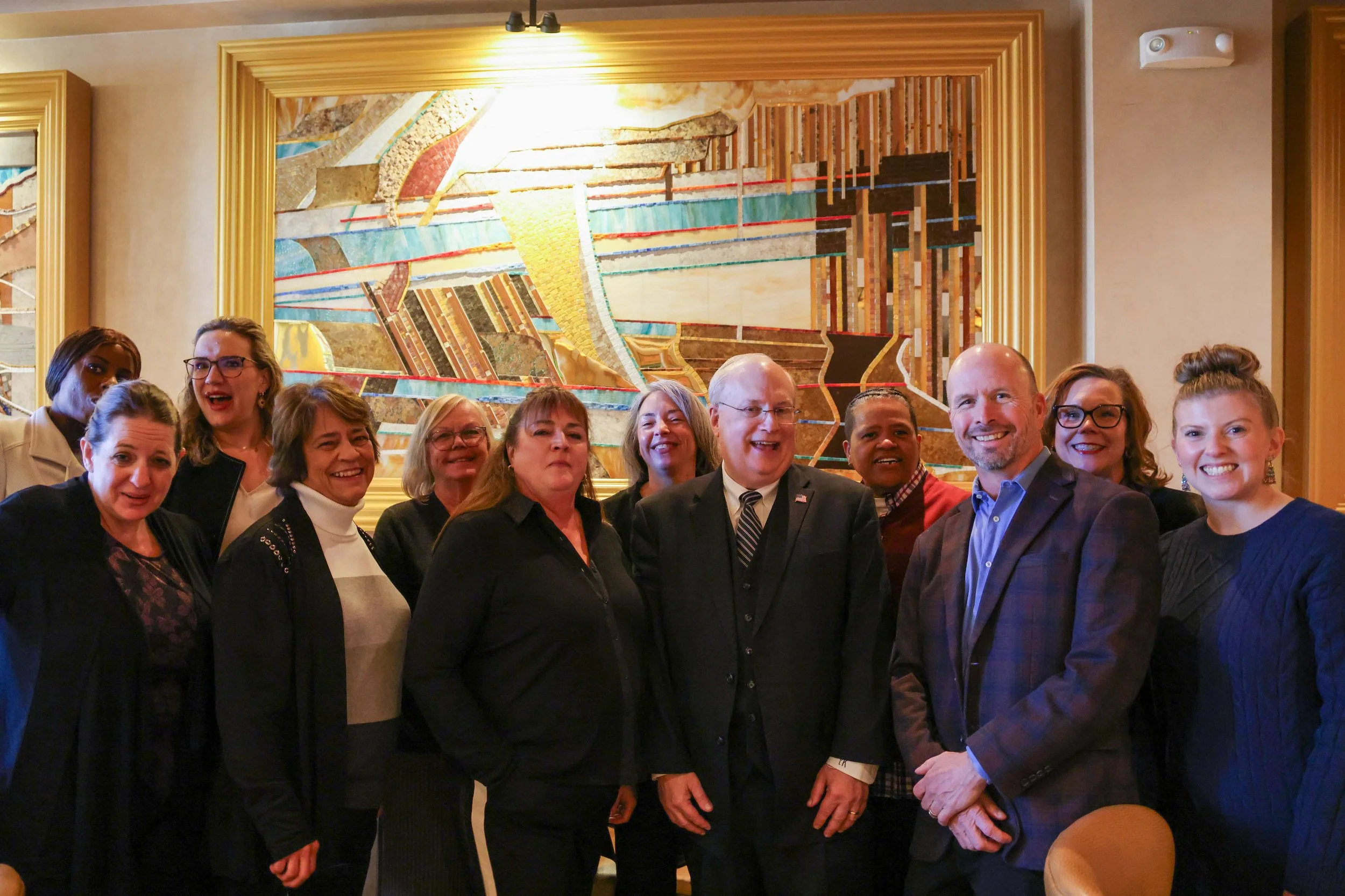 A group of people standing together indoors in front of an abstract artwork with a golden frame.
