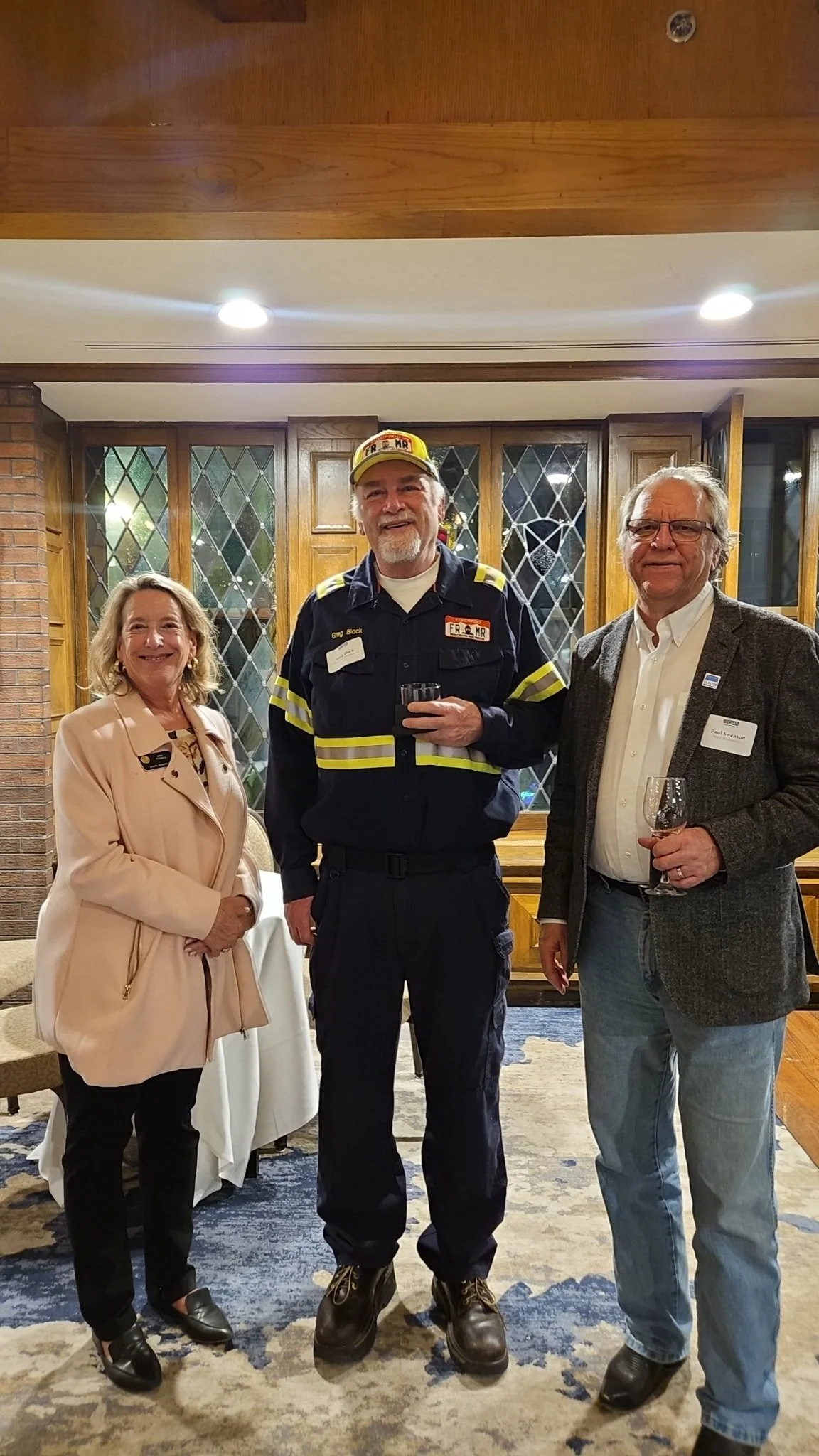 Three people standing together indoors at a formal event, smiling and holding glasses, with a woman on the left wearing a beige coat, a man in the middle dressed as a firefighter, and a man on the right in a blazer with a wine glass.