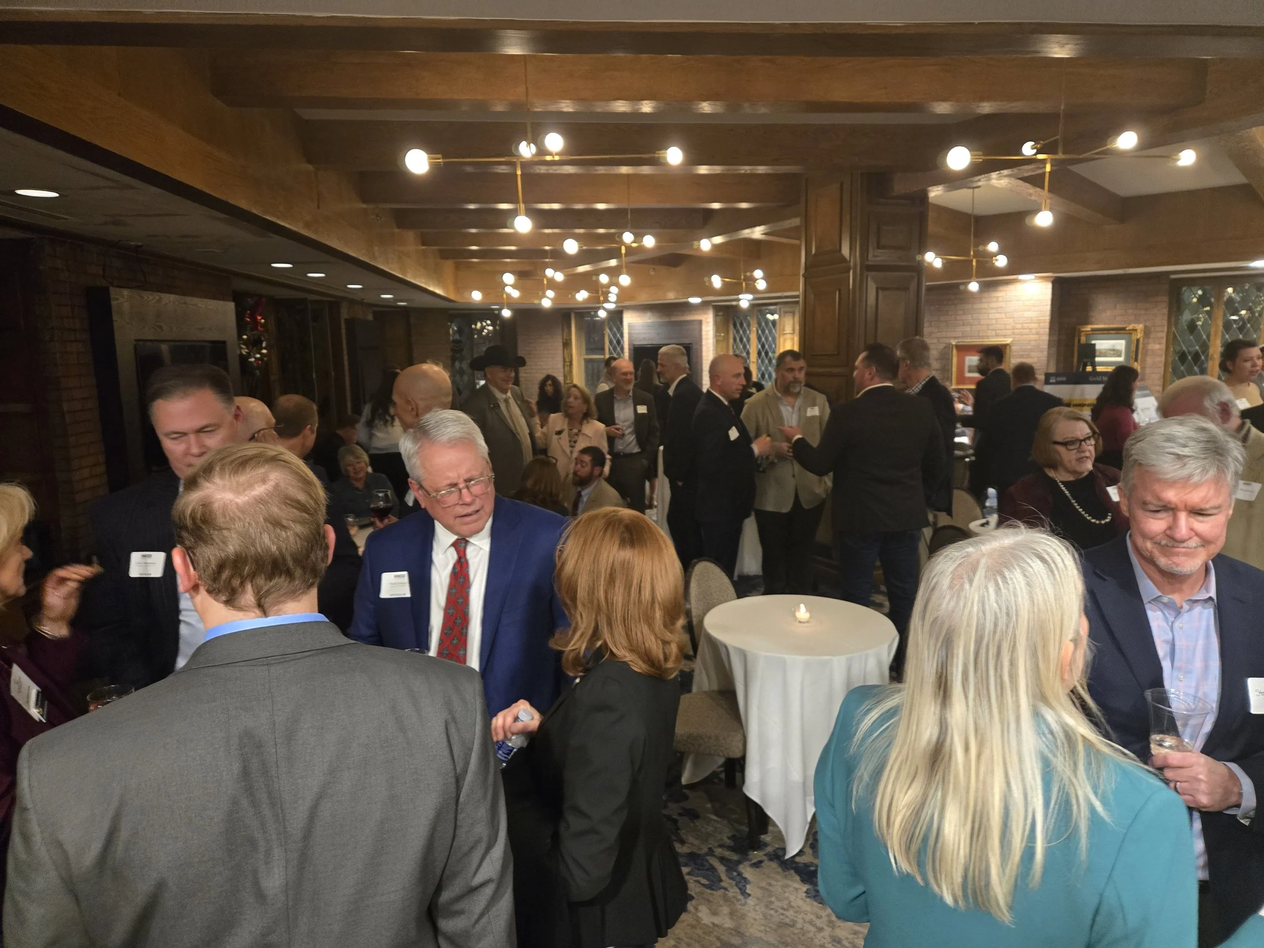 A group of people at a formal networking event or reception, standing and conversing in a warmly lit, wood-paneled room with brick walls and framed pictures.