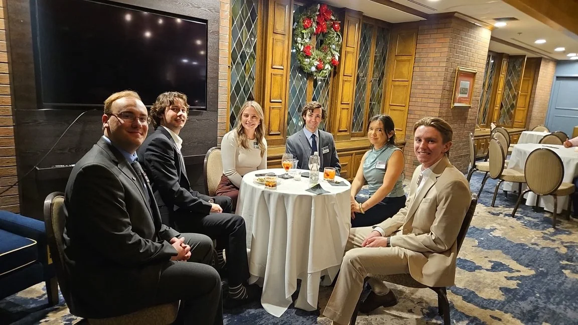 Six people sitting around a round table at a formal event, with Christmas decorations on the wall in a decorated banquet hall.