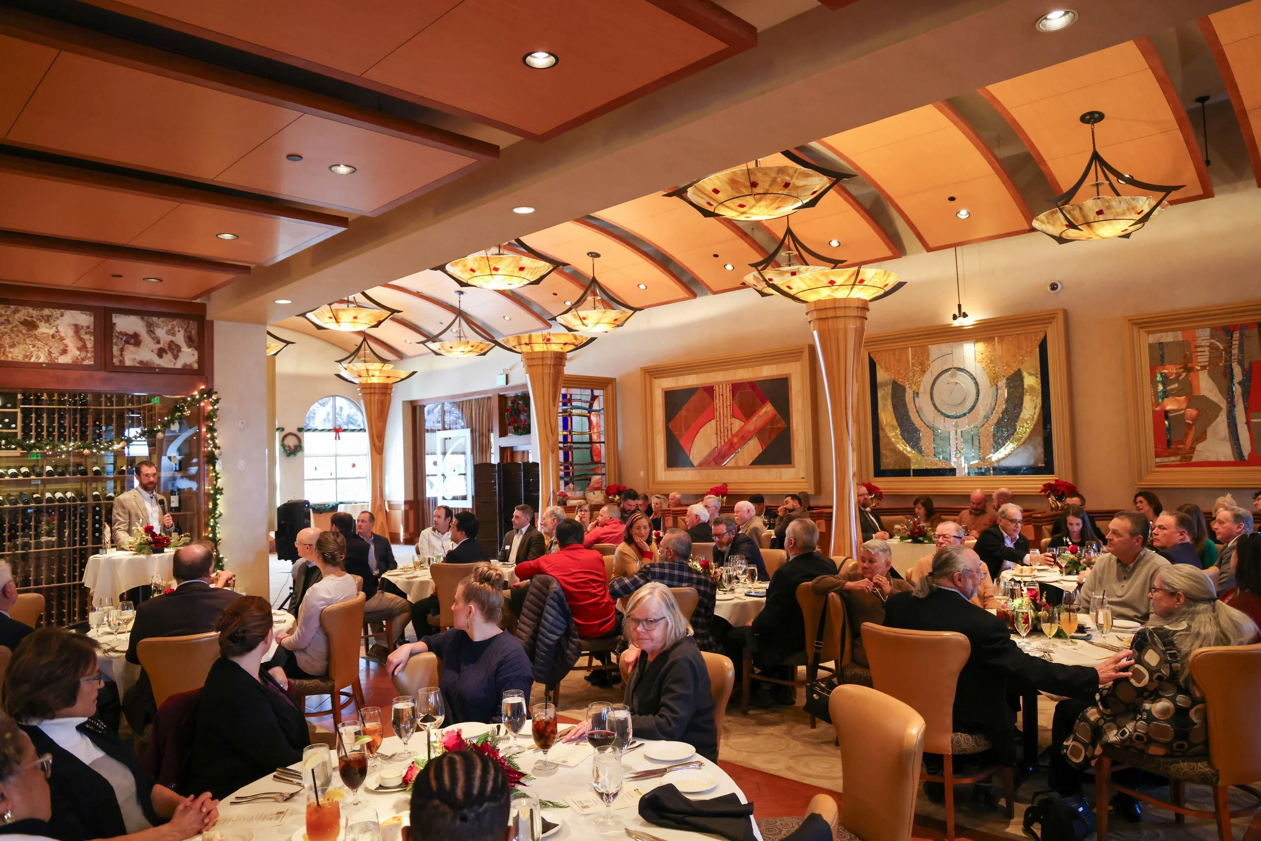 A restaurant or banquet hall filled with people seated at tables, decorated with flowers and glasses, with a man standing and speaking at a small podium. The ceiling has warm lighting fixtures, and windows with holiday wreath decorations are visible 