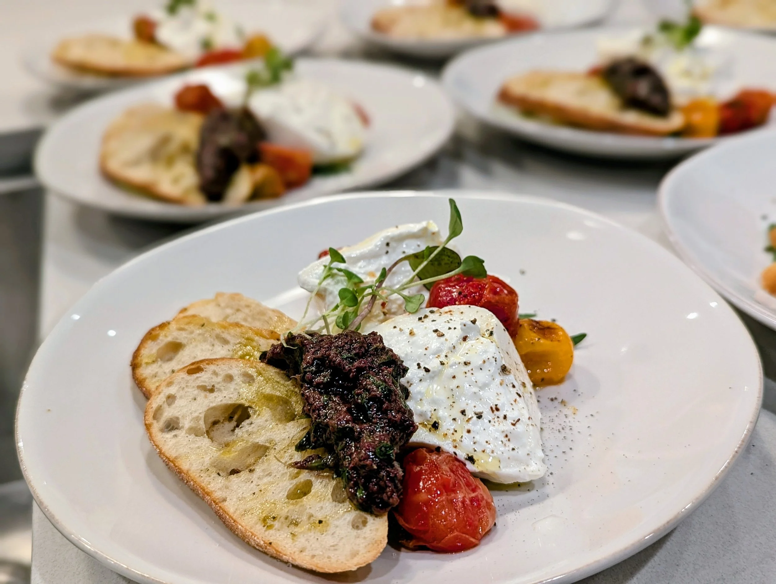 Close-up of a plate with a slice of bread topped with toppings including a dollop of white creamy sauce, cherry tomatoes, greens, and a dark tapenade or spread, with multiple similar plates in the background.