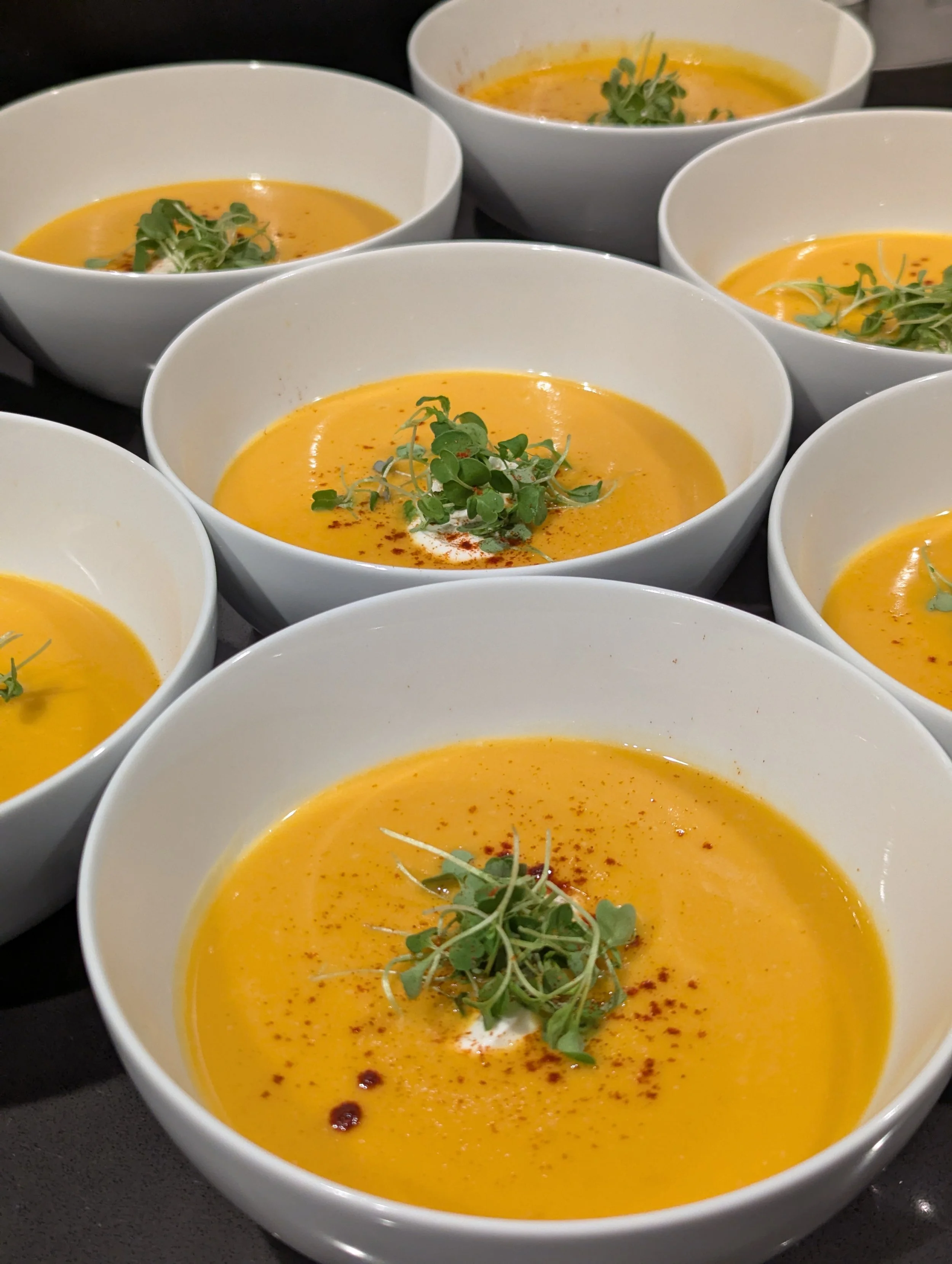 Several white bowls filled with creamy orange soup garnished with microgreens and sprinkled with spices on a dark surface.