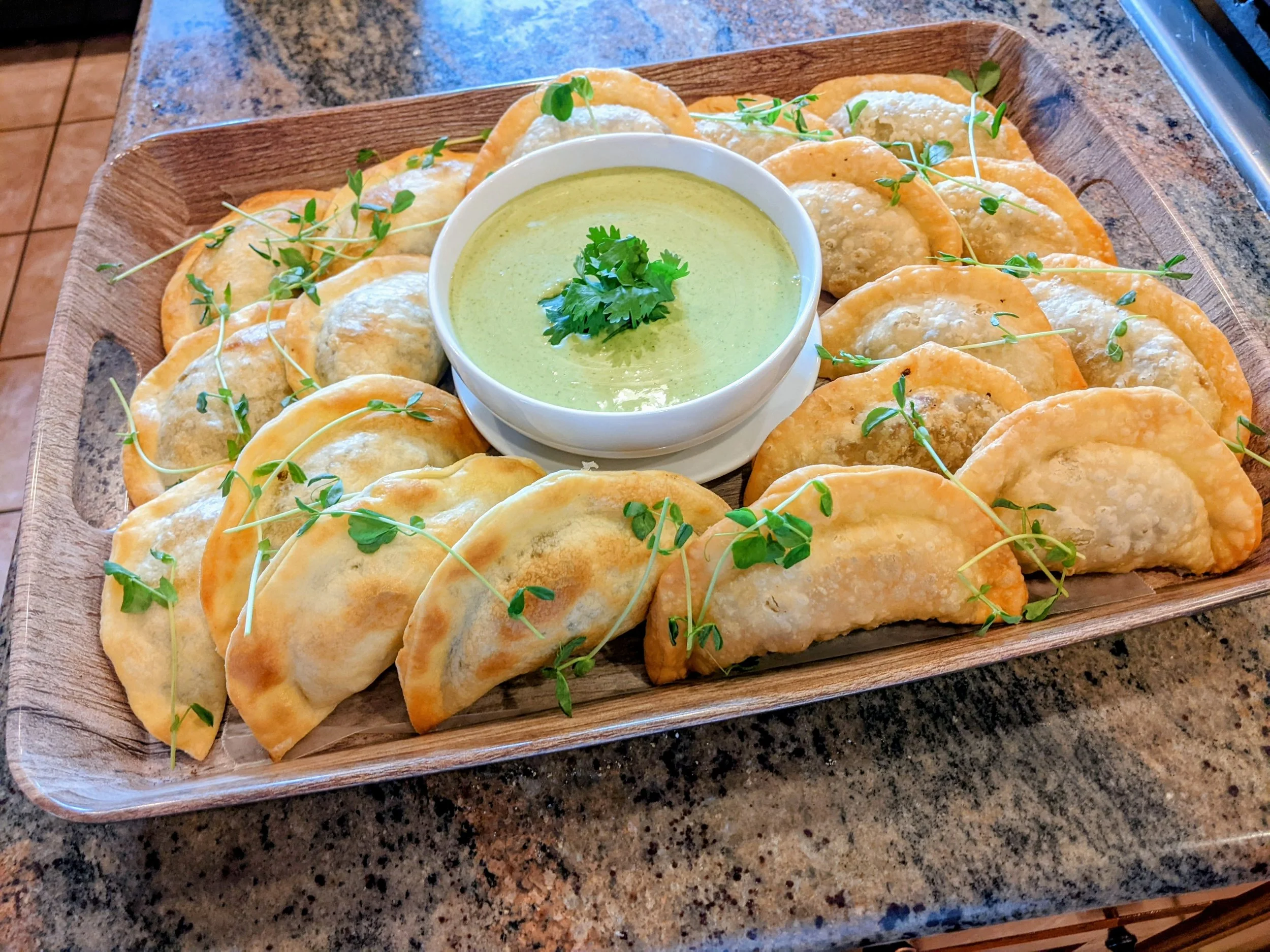 A wooden tray with fried empanadas garnished with microgreens, served with a bowl of green dipping sauce garnished with cilantro.