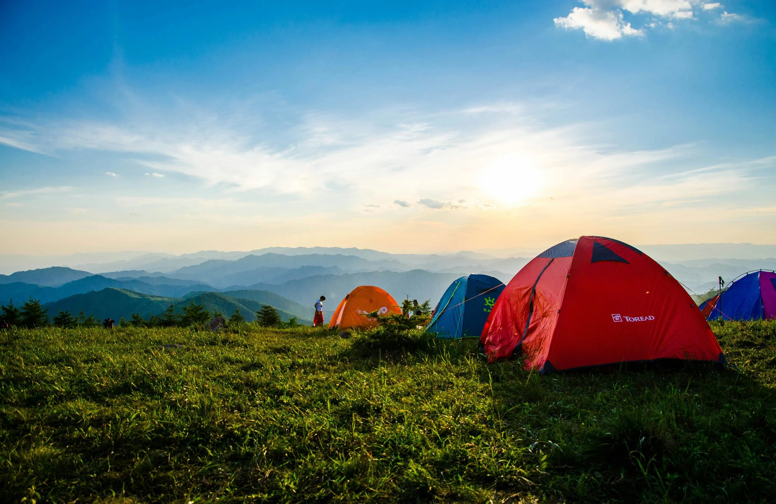 Multiple colorful tents set up on a grassy mountain meadow with distant mountain ranges under a partly cloudy sky during sunset.