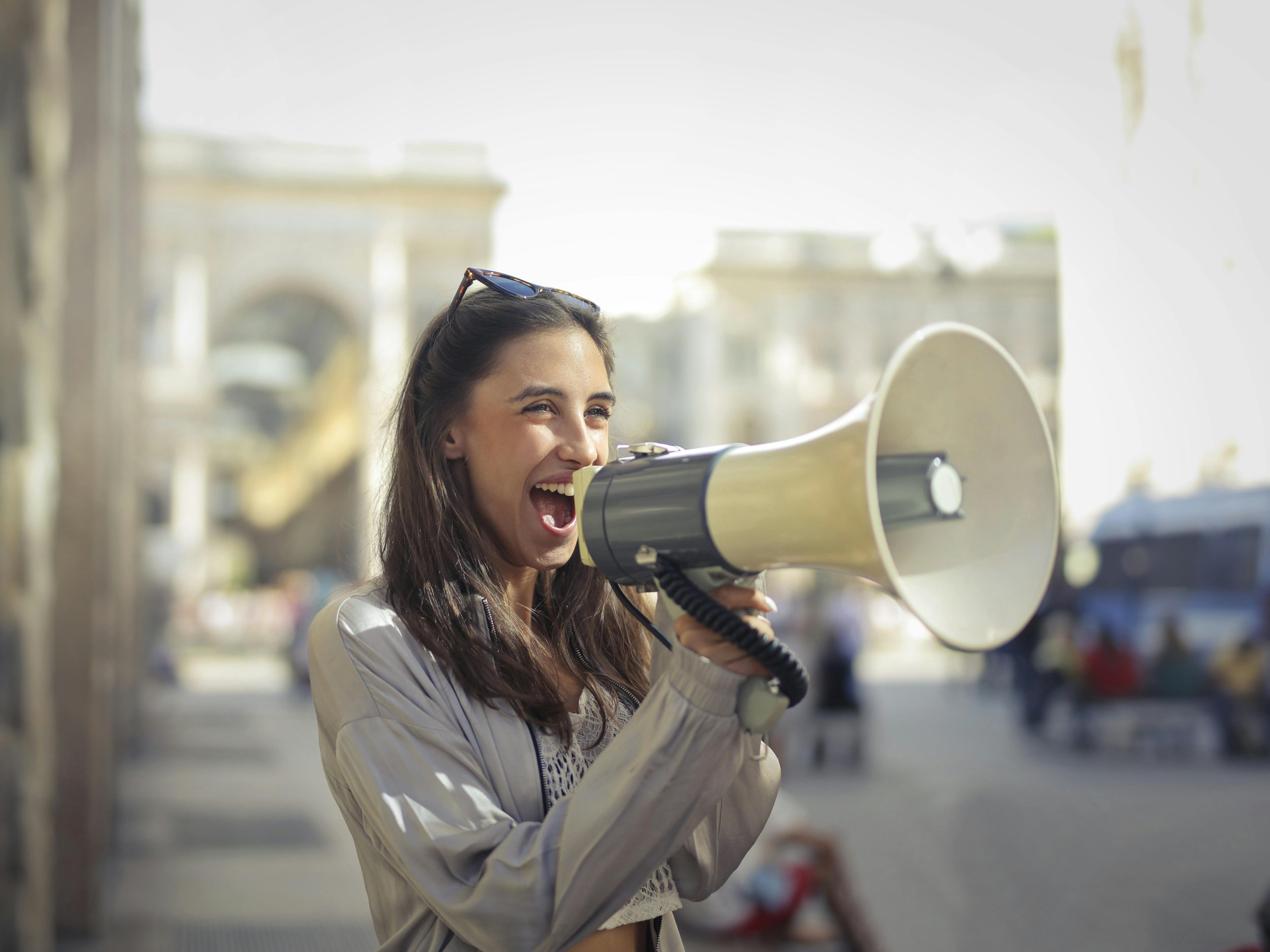 A woman outdoors using a megaphone, smiling and shouting.