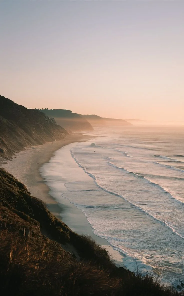 Sunset over a beach with cliffs, gentle waves, and a pink sky.