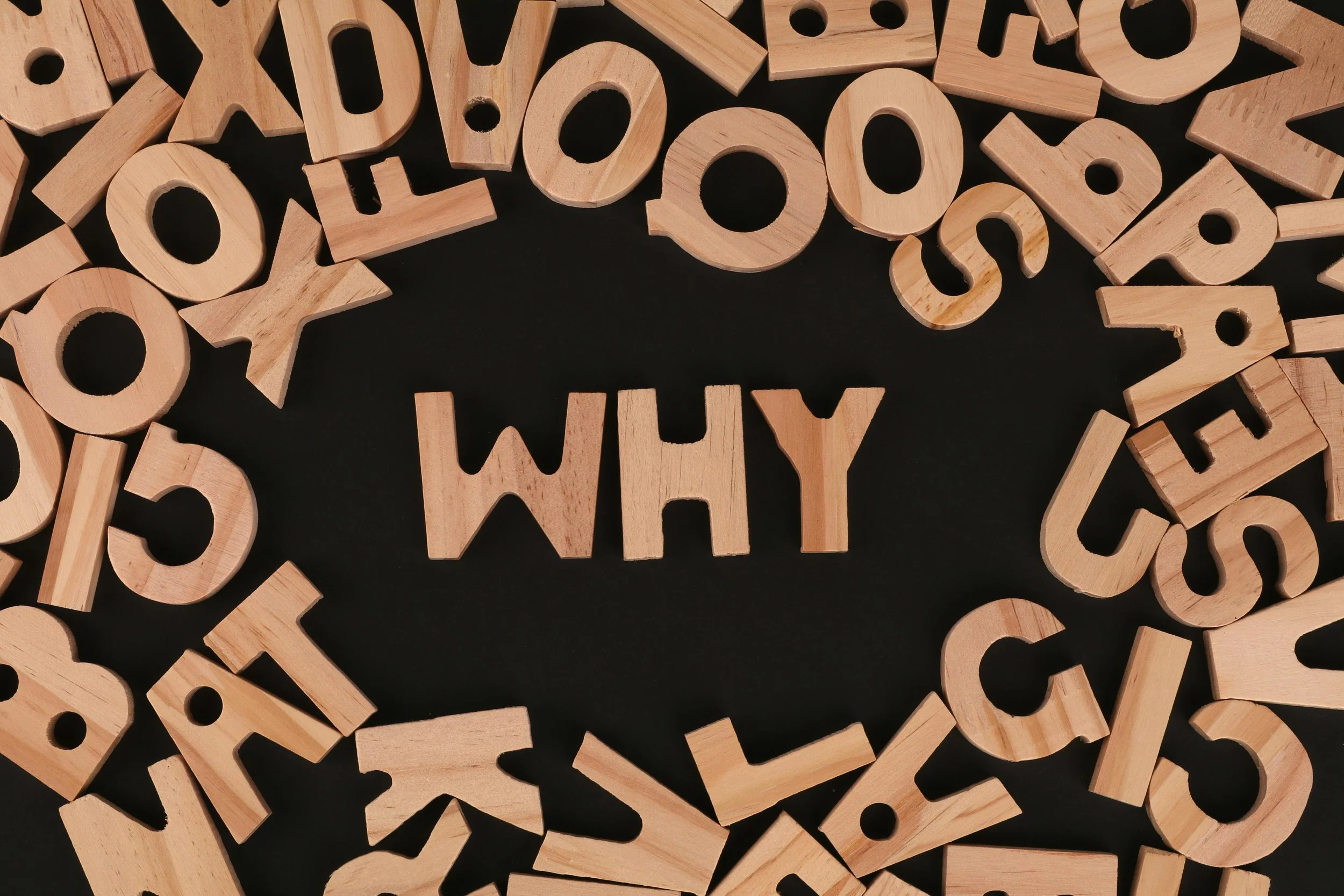 Wooden letter cutouts scattered on a black surface with the word 'WHY' spelled out in the center.