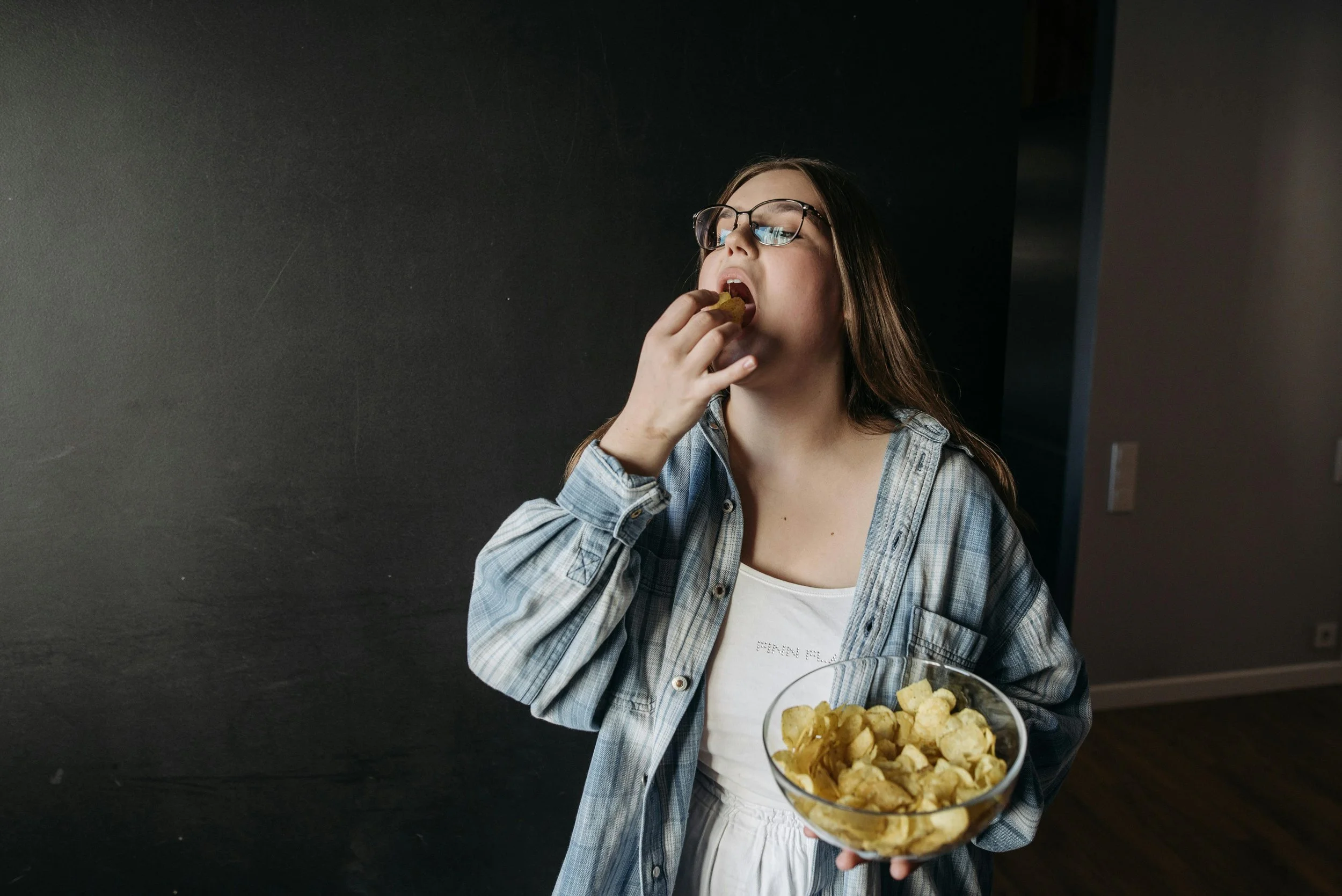 A young woman with glasses, wearing a plaid shirt and white top, is eating potato chips from a glass bowl.