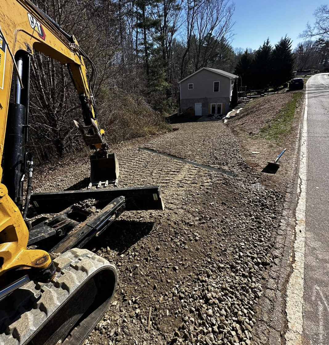 Construction vehicle with digging arm on a dirt and gravel patch beside a road, with a house and trees in the background.