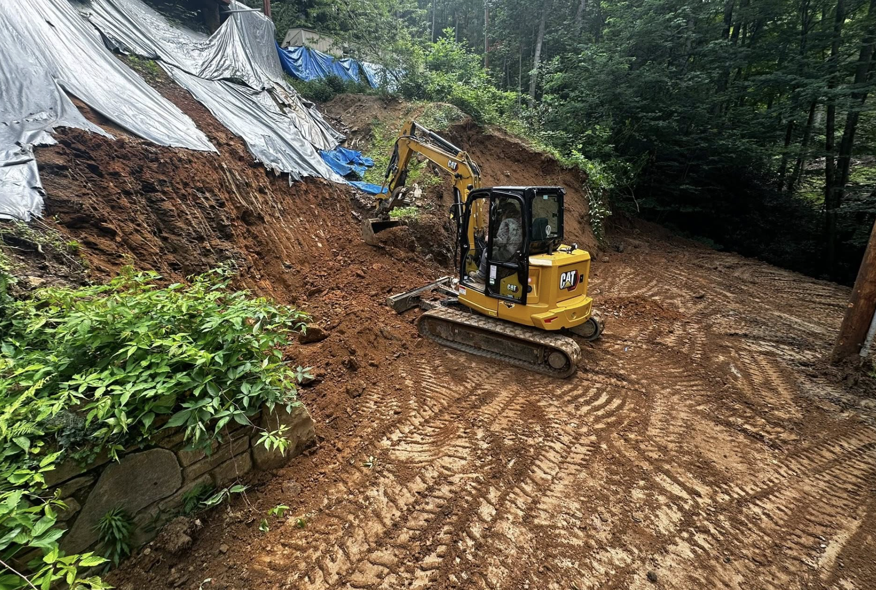Construction site with a yellow mini excavator digging into the earth, with trees and foliage on a hillside, some tarp covering the slope, and tire tracks in the dirt.