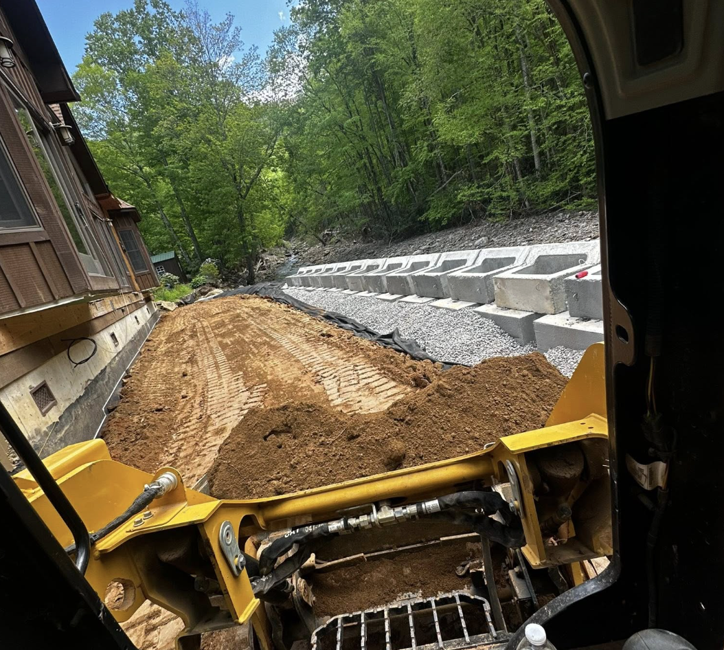 View from inside a construction vehicle, looking out onto a dirt and gravel path flanked by a house on the left and a forested hillside on the right, with barriers along the path.