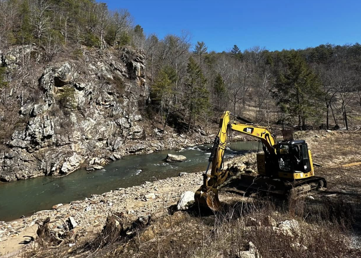 A yellow and black excavator working near a river with rocky banks, surrounded by a wooded hillside and a clear blue sky.