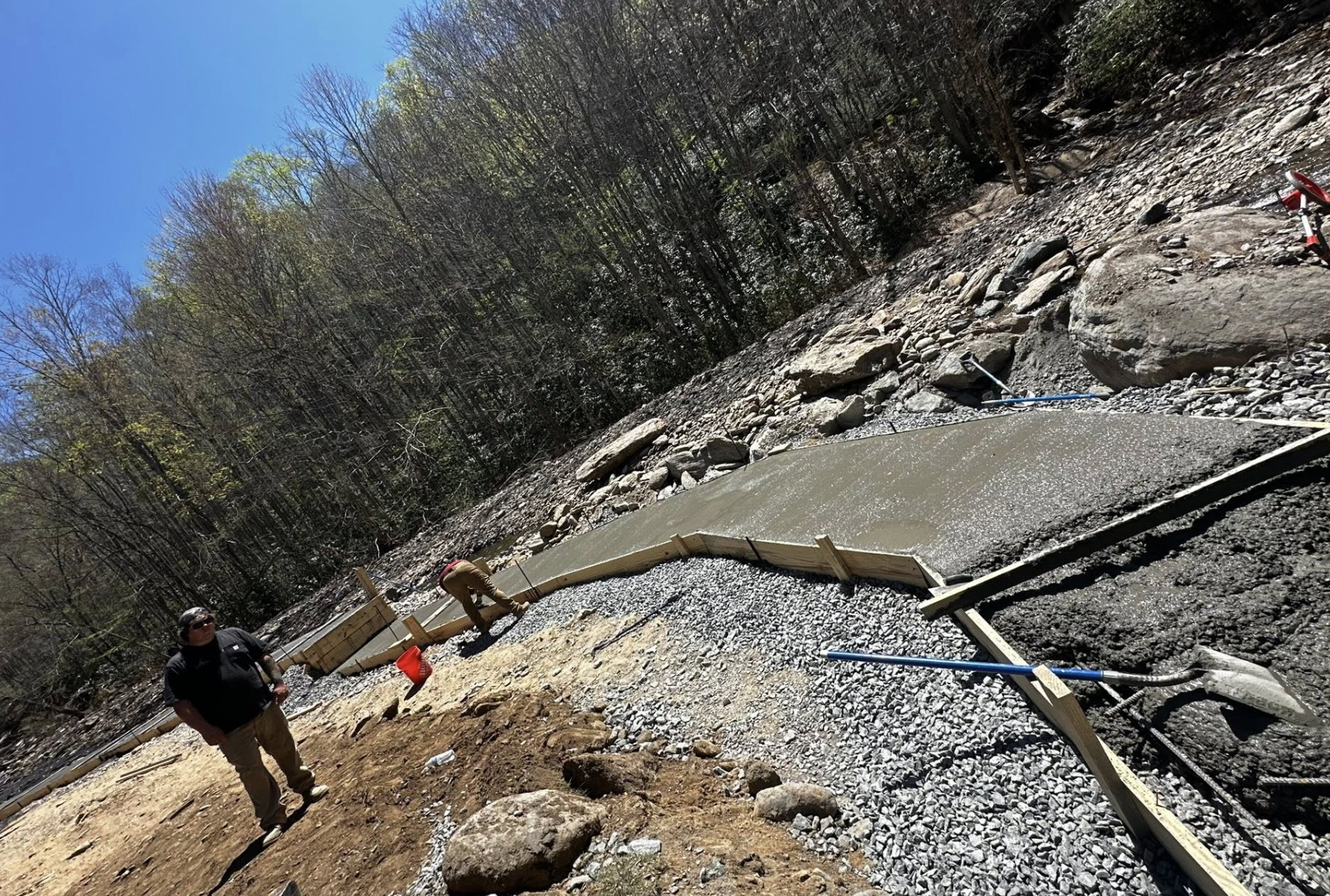 Construction workers building a concrete sidewalk along a rocky hillside, with some workers pouring and finishing concrete, and construction tools visible.