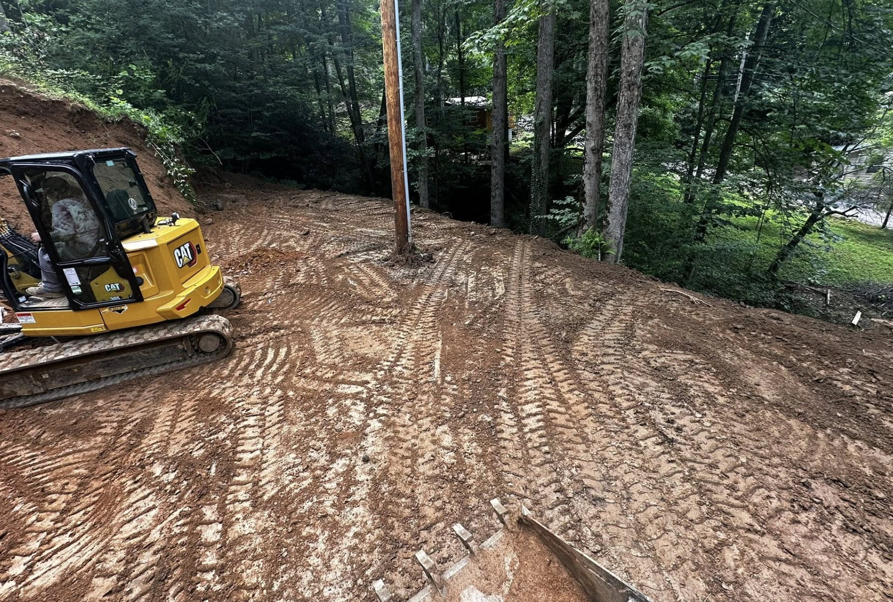 Construction site with dirt and tire tracks, a small excavator on the left, and trees in the background.