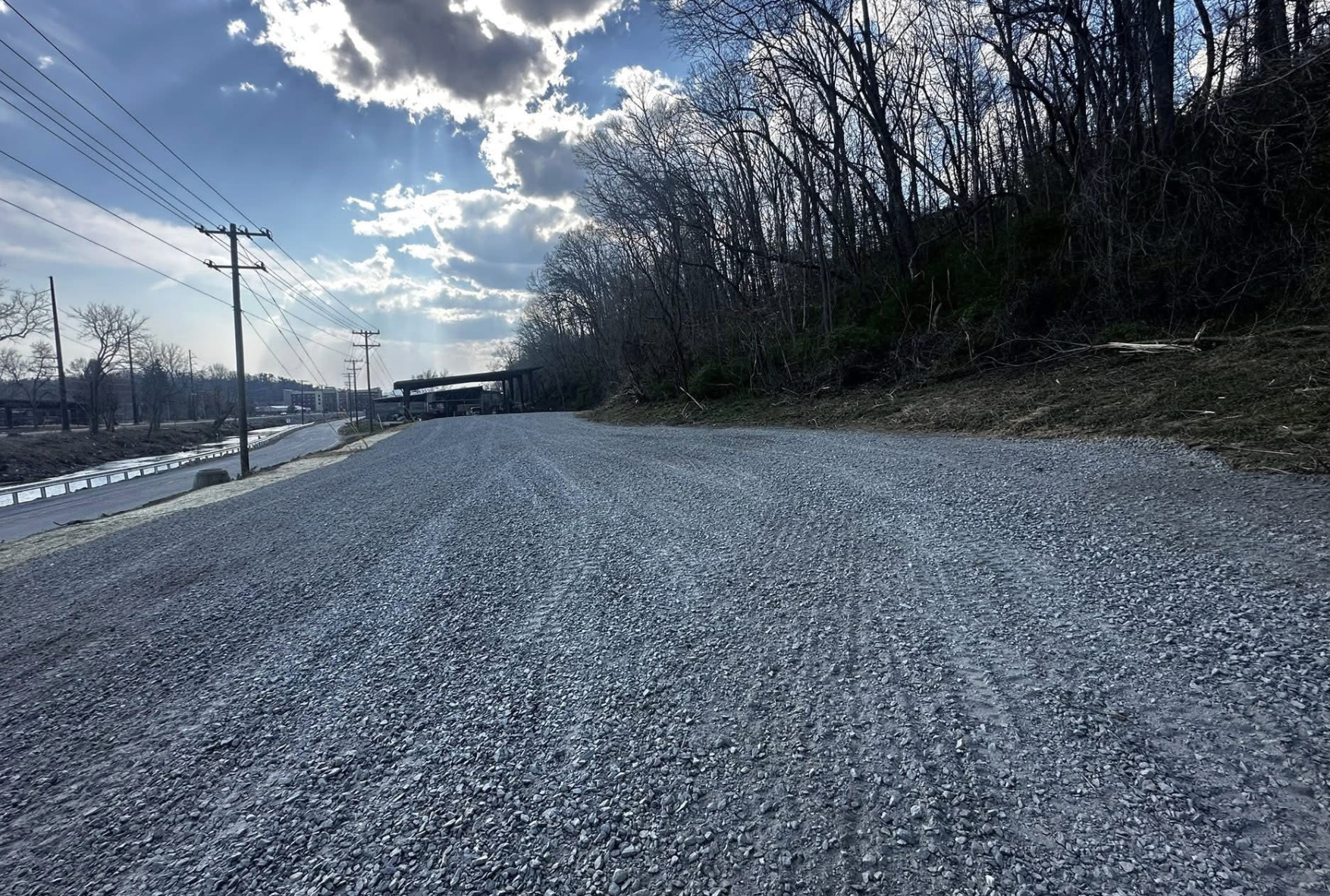 A gravel road running alongside a hill with leafless trees and utility poles, under a partly cloudy sky with sunlight streaming through clouds.