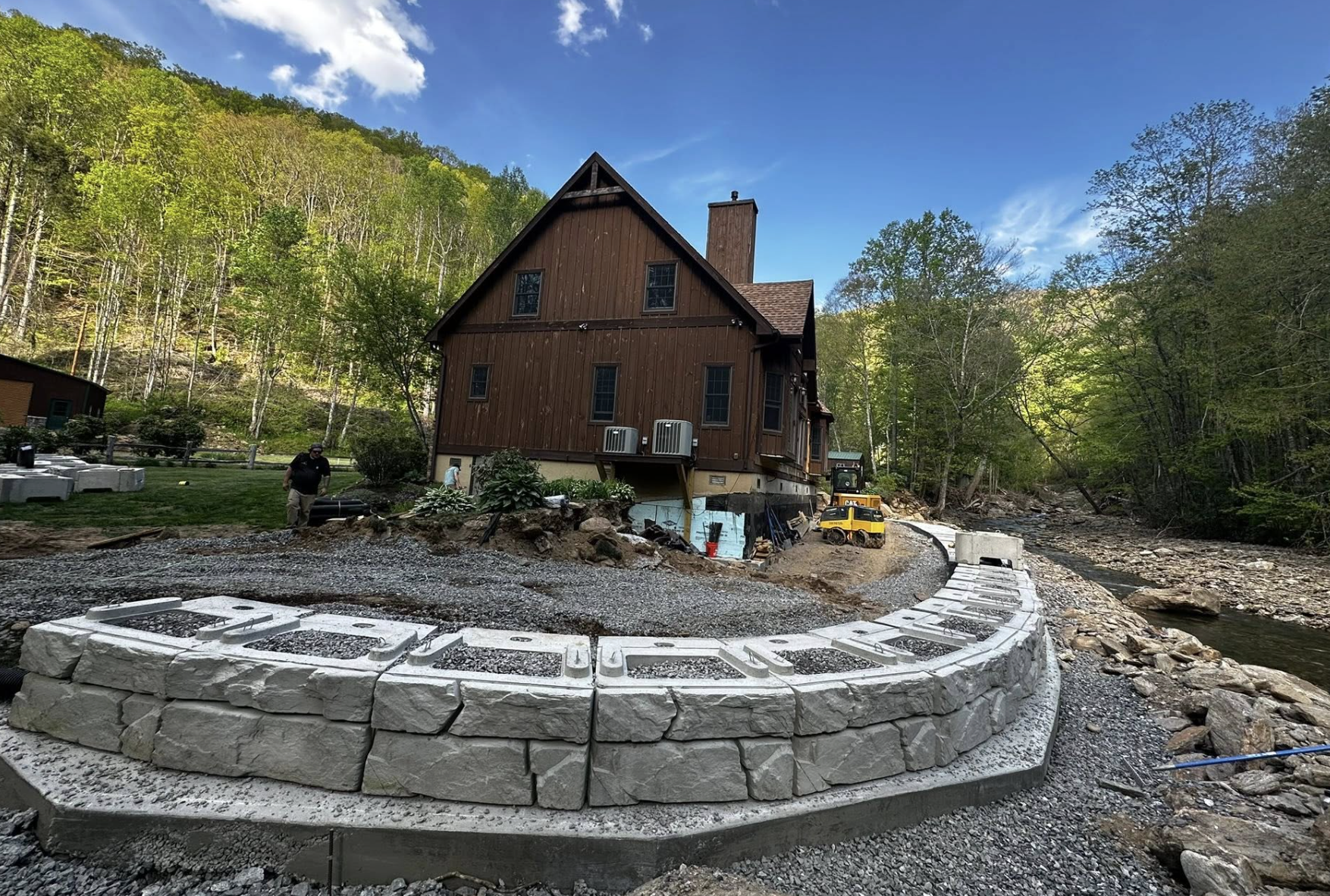 A house in a wooded area under construction with a curved stone retaining wall and construction equipment nearby.