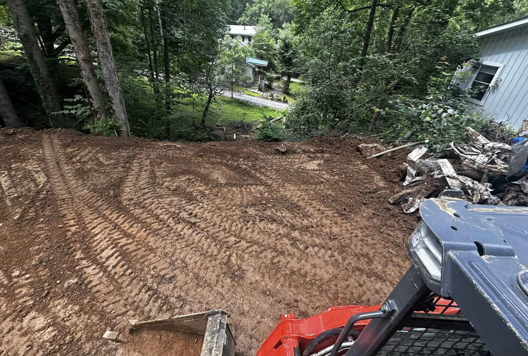 A cleared construction or landscaping site with brown dirt and tire tracks, surrounded by trees and residential houses in the background.