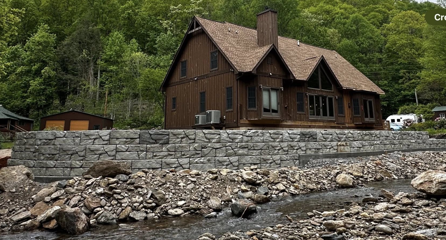 A wooden house with a steep roof and multiple windows, located on a rocky riverbank with a stone retaining wall, against a backdrop of lush green forest.
