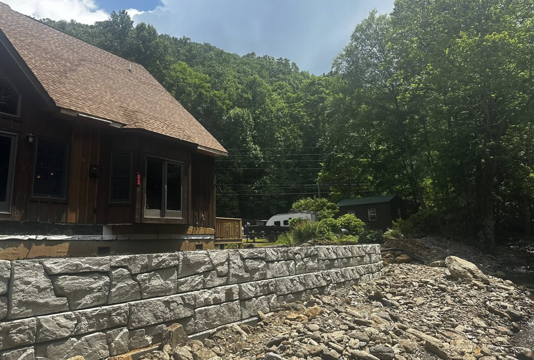 A house built on a stone retaining wall next to a rocky riverbank, with dense green trees and hills in the background under a partly cloudy sky.