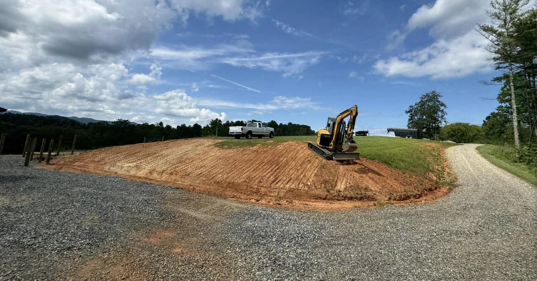 Construction site with a small yellow excavator on a dirt hill, a gravel road in the foreground, a white pickup truck on the hill, and a gravel driveway on the right, with a backdrop of trees and a partly cloudy sky.