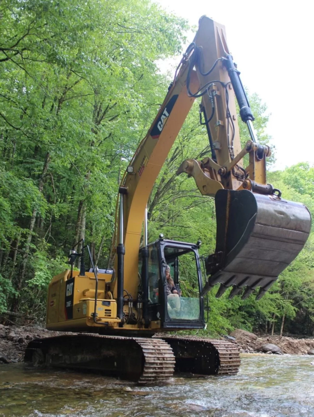 A yellow Caterpillar excavator with a large bucket attachment, operating in a forested area with green leafy trees, near a shallow river or stream.