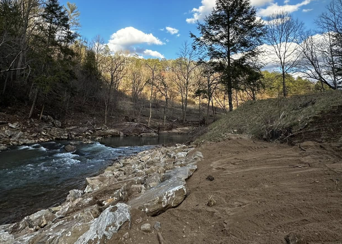A river flowing through a wooded area with a dirt path on the right side, lined with rocks and leafless trees under a partly cloudy blue sky.