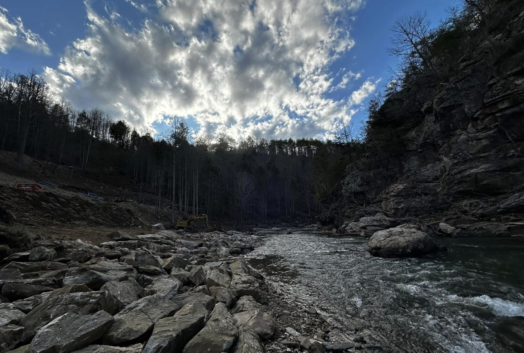 A river flowing through a rocky landscape with leafless trees on the hillside, under a partly cloudy sky.