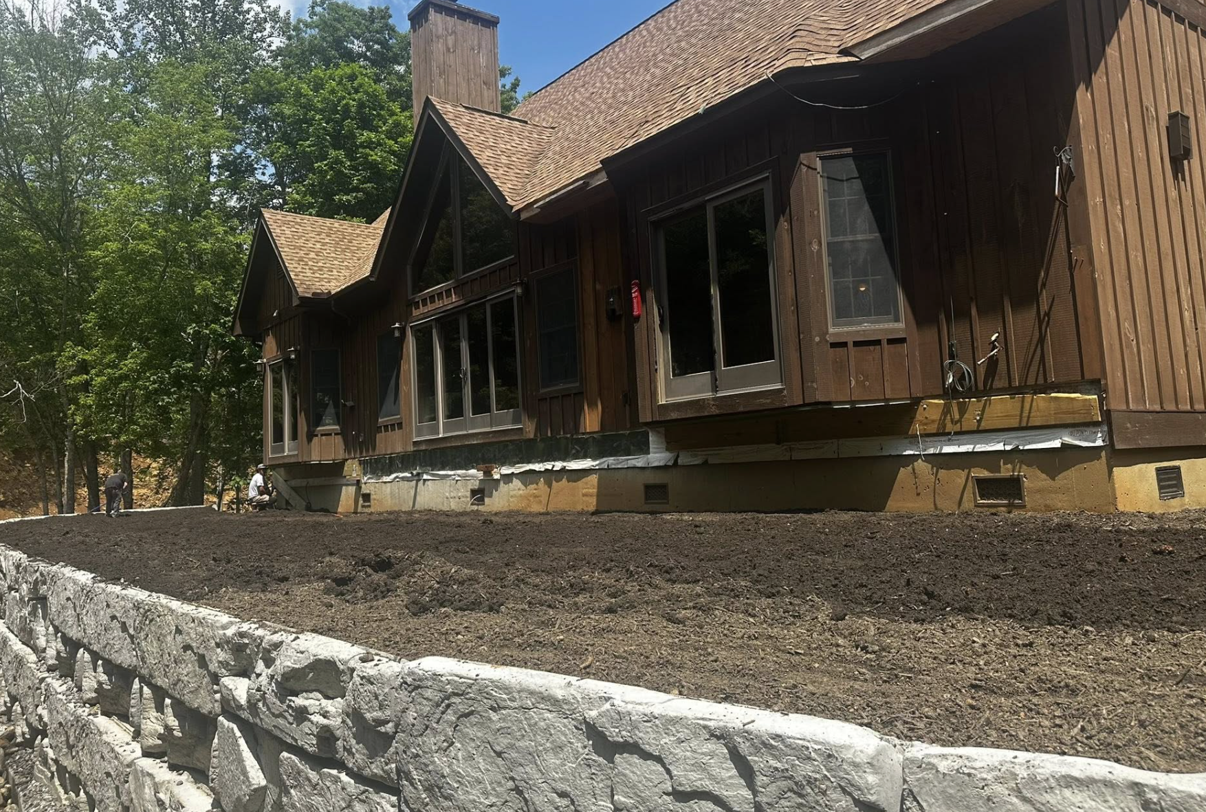 A house under construction with a wooden exterior, large windows, and a sloped roof, surrounded by trees. The ground around the house is bare soil, with a stone retaining wall in the foreground.