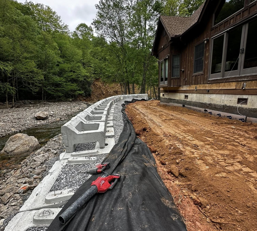 Construction site next to a wooden house with a curved retaining wall being built, gravel and dirt ground, trees in the background, construction tools and materials present.