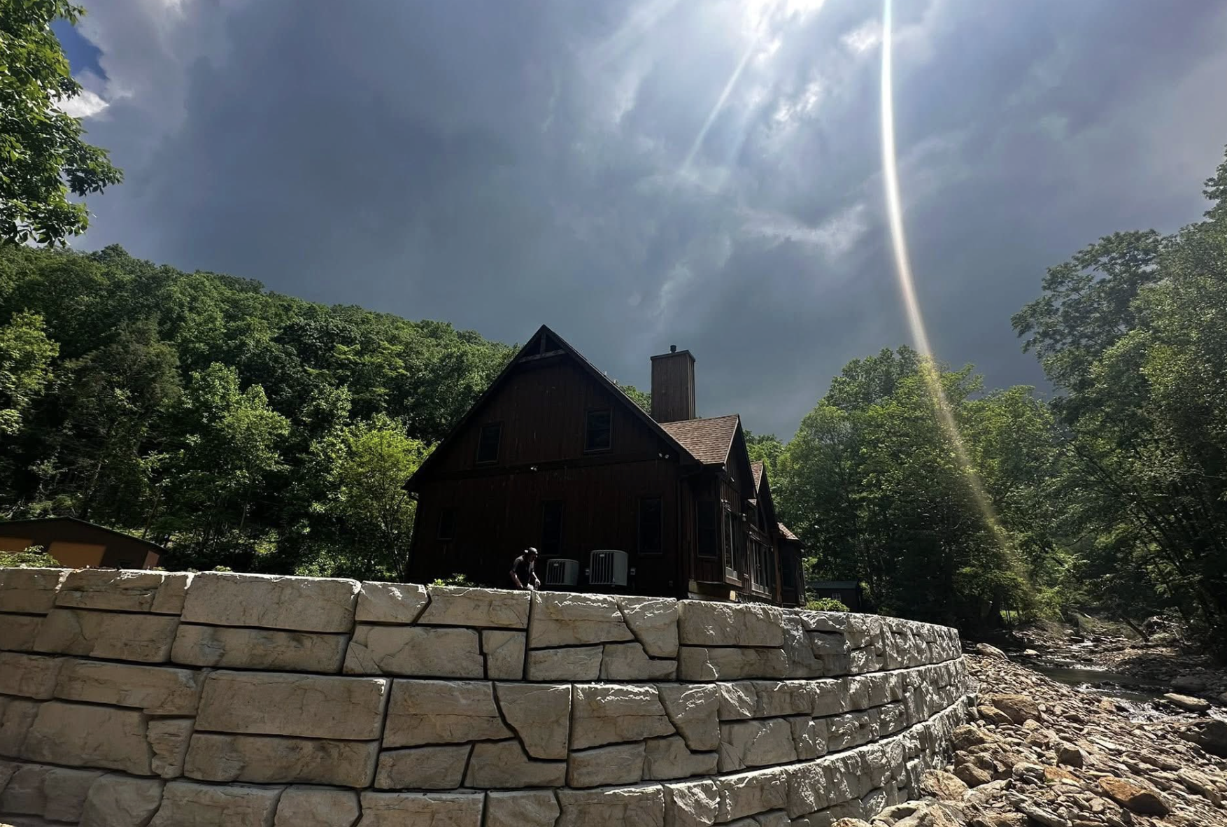A dark wooden house or cabin on a raised stone foundation, surrounded by lush green trees, under a partly cloudy sky with sun rays.