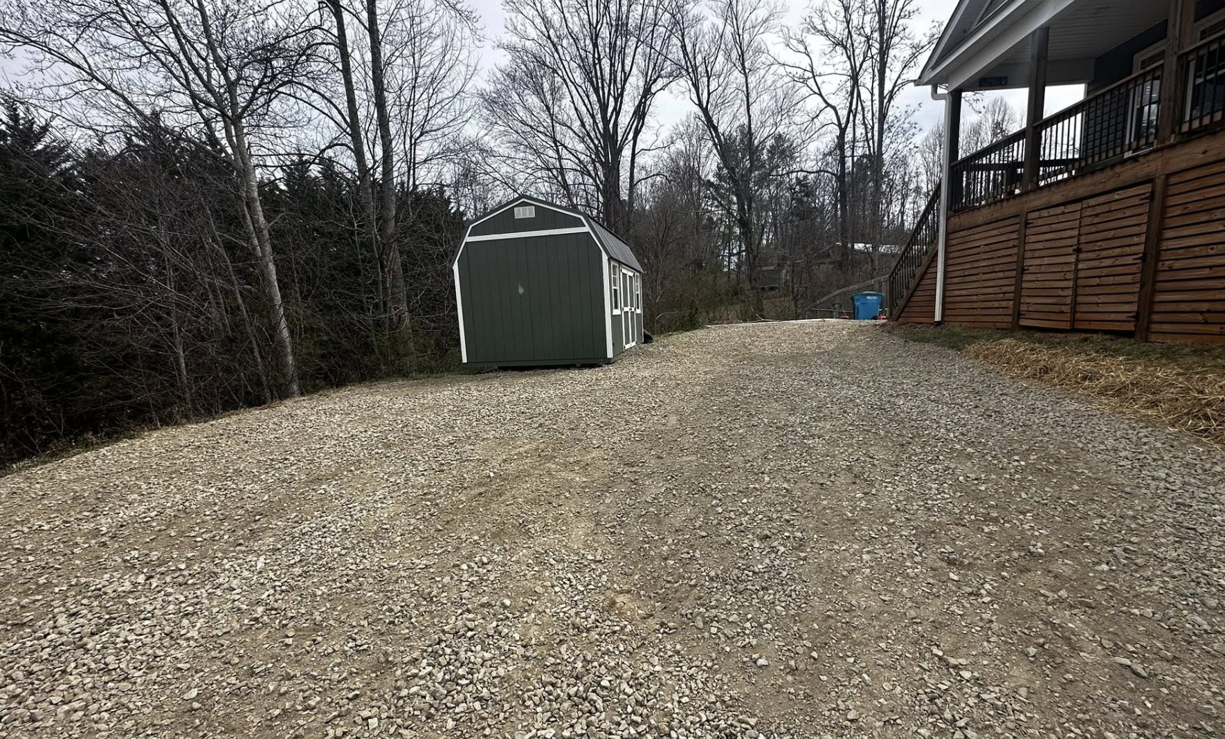 Gravel driveway with a green shed and a wooden fence beside a house, leafless trees in the background, overcast sky.