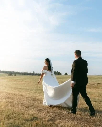 Bride and groom take their wedding photo amongst green landscape while the groom raises the bride's white wedding dress tail.