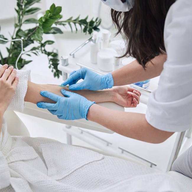 Nurse with dark hair administering an infusion in a clinic space with green plant.