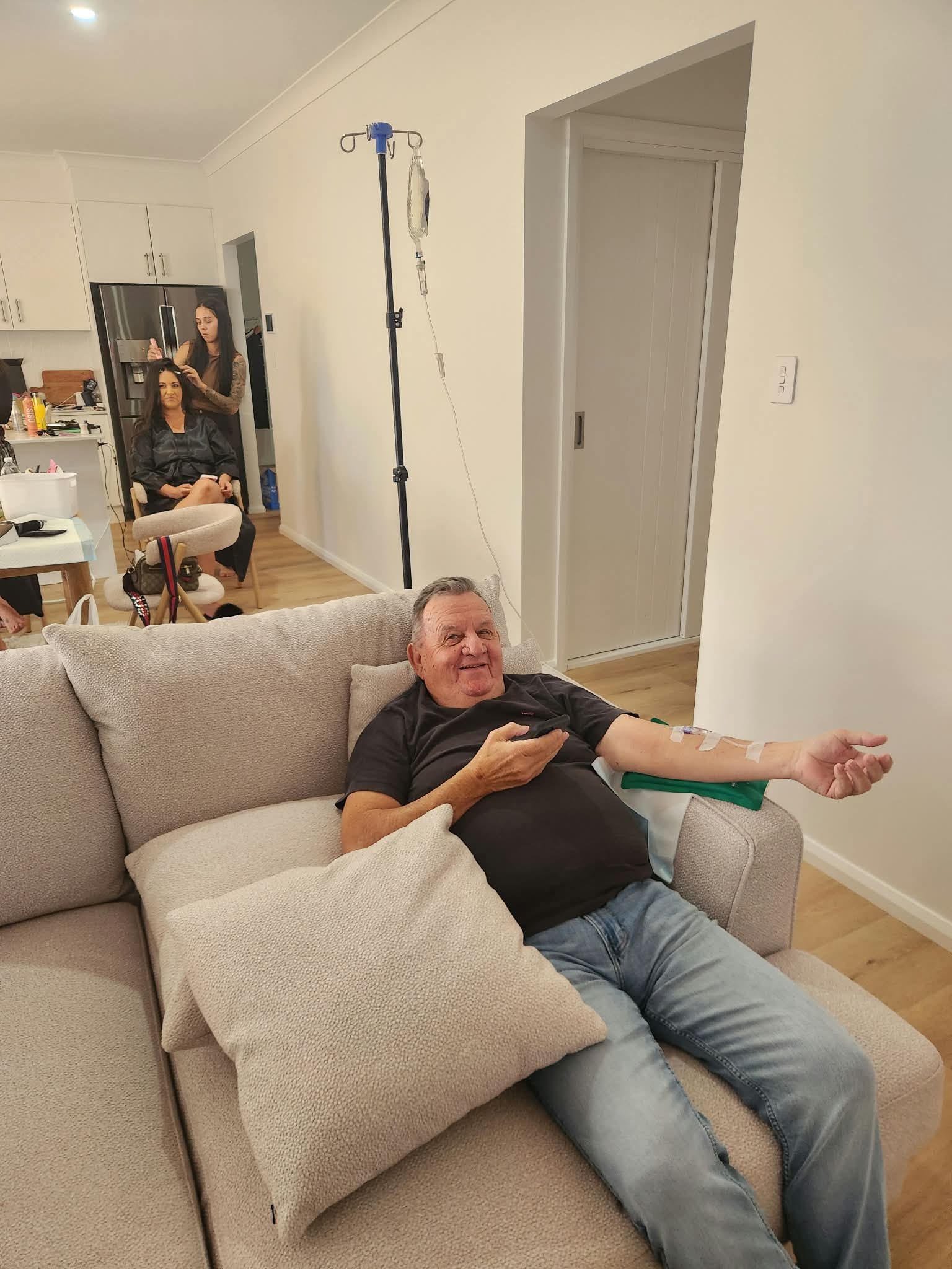 A mature man sits comfortably on a large couch with arm out and IV therapy infusions set up. A woman is in the background getting her hair done in preparation of the wedding party.
