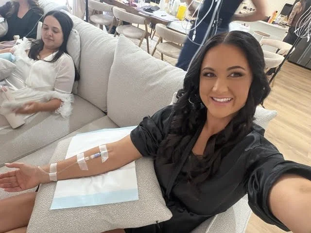 A girl with dark long hair is relaxing on a lounge with IV Therapy injections shown in her arm as she receives Infinity Infusions wellness therapy. Another woman is sitting next her relaxing as she completes her treatment for the wedding booking.