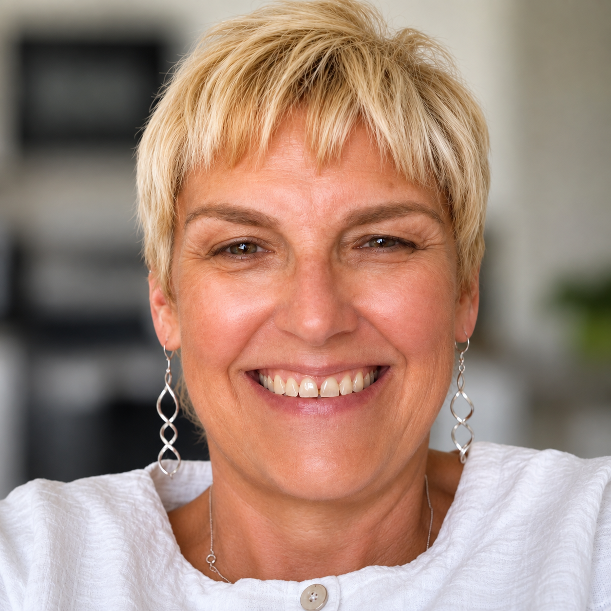 Close-up of a smiling middle-aged woman with short blonde hair, wearing earrings and a white top.