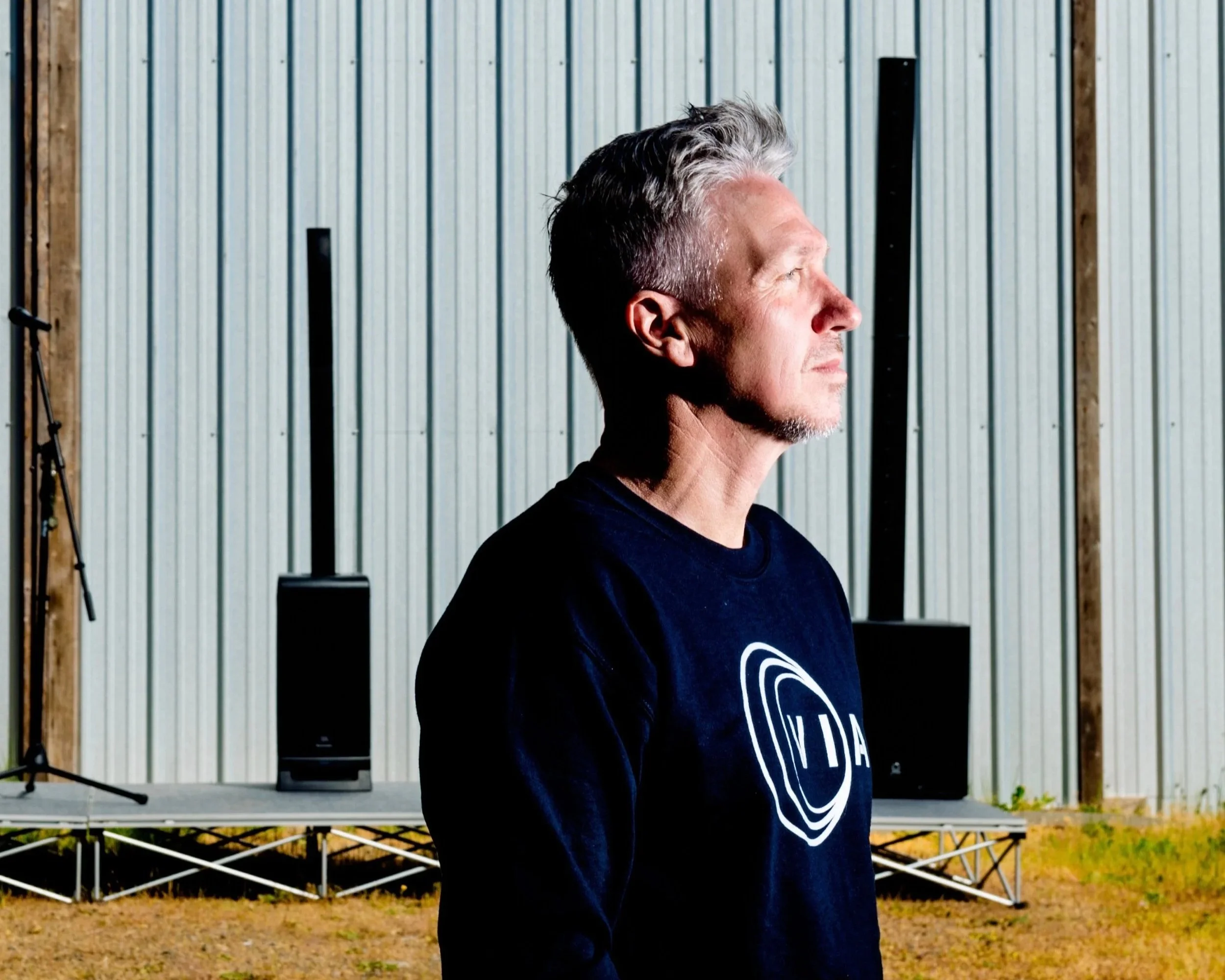 Rob Bowker stands outdoors facing sunlight with a metal building and outdoor stage equipment in the background.